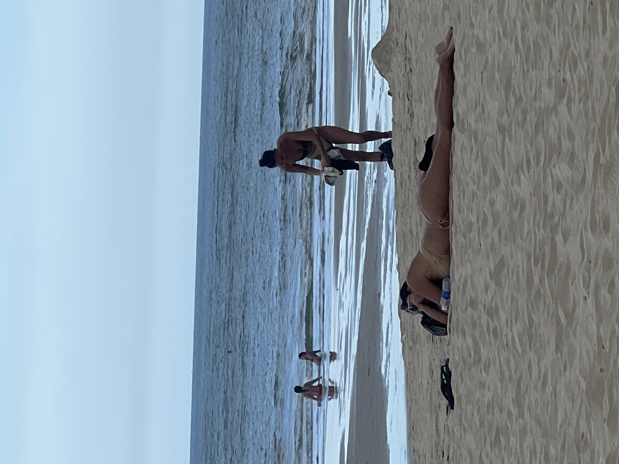 This image depicts a beach scene in Đà Nẵng, Vietnam. In the foreground, a person is lying on their stomach on the sand, facing away from the camera. They are wearing a light-colored bikini and sunglasses, with a water bottle and a bag next to them. Further back, a person is standing on the wet sand near the water's edge, holding a dark garment and a white cloth. They are wearing a dark bikini. In the water, two other individuals are visible. One person is sitting in the shallow water, and another is wading further out. The ocean is calm with small waves, and the sky is overcast. The beach is sandy with a gentle slope towards the water.