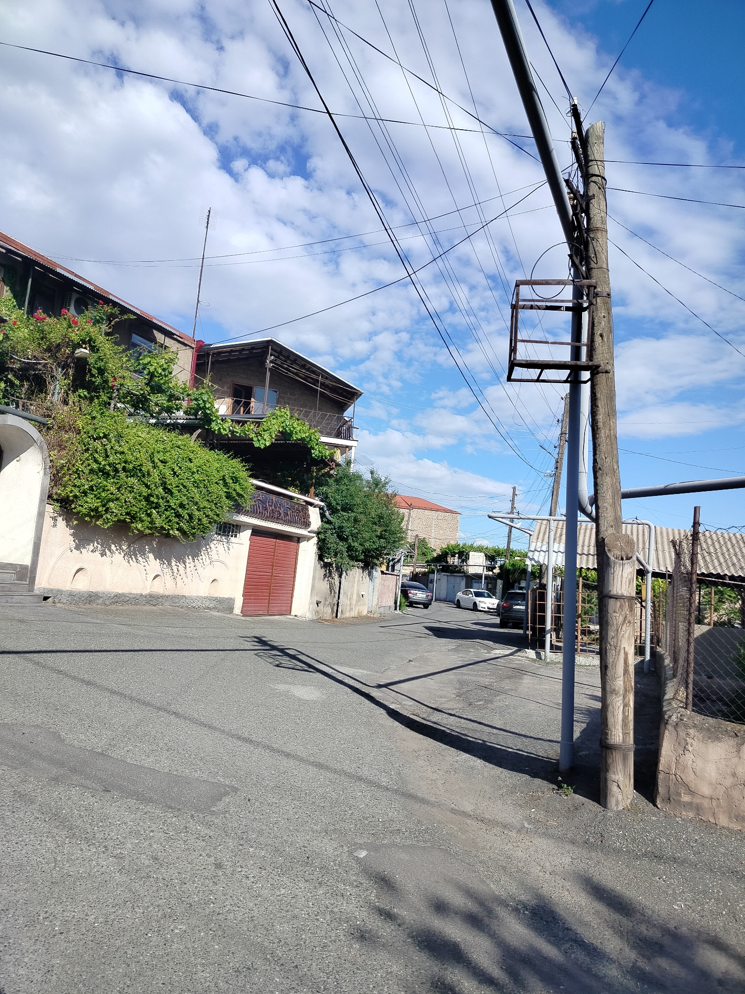 A street scene in Yerevan, Armenia. The scene is during the daytime with a partly cloudy sky overhead. A utility pole stands prominently in the right foreground, with a metal fixture attached partway up. Overhead, numerous electrical wires crisscross the sky.
To the left is a building partially covered in greenery, with a red garage door. A second story has a balcony. Across the street, parked cars are visible, along with a fence, a shed, and other houses. The road is paved with asphalt and shows signs of wear and repair. Shadows from the utility pole are cast across the street. There are no people visible in the scene.