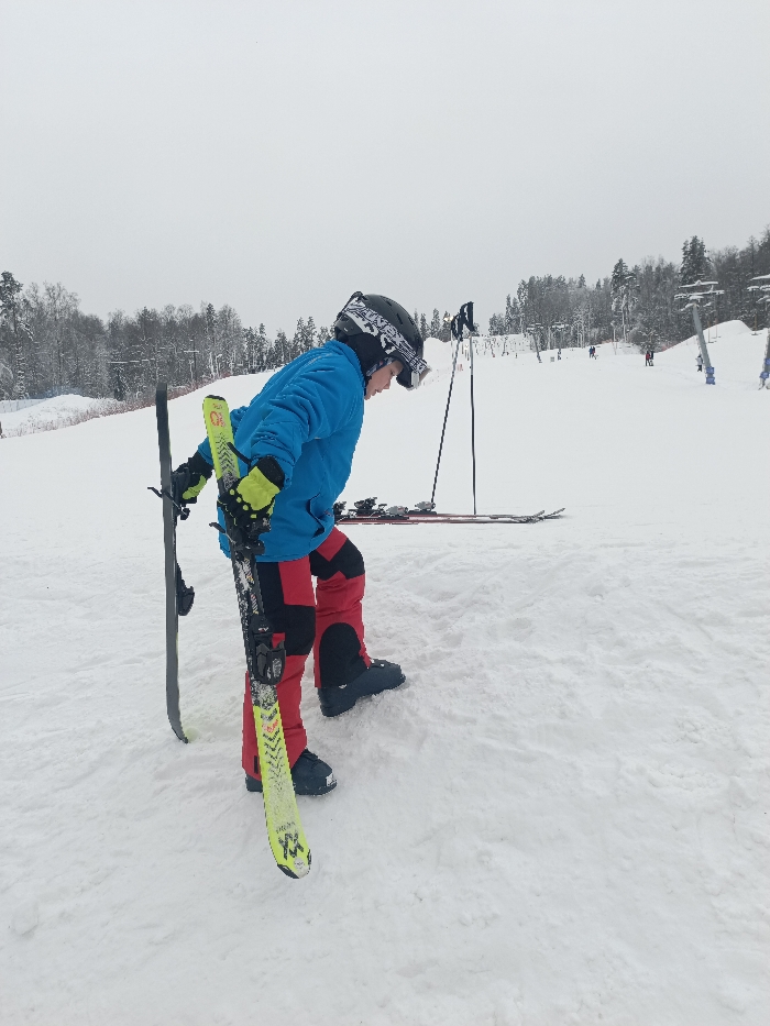 A ski resort scene in Svetloe, Russia, depicts a person, likely a child or adolescent, standing on a snow-covered slope under an overcast sky. The individual wears a blue ski jacket, red and black ski pants, black ski boots, yellow-green gloves, and a black ski helmet with goggles resting on top. They hold two skis, one notably yellow-green with black markings and the other dark grey, vertically in their left hand. Their body is angled to the right, with their head slightly lowered and right foot partially lifted. In the immediate background, a second pair of skis and ski poles are laid flat on the snow. The broader environment shows a wide, white ski run, bordered by snow-covered coniferous trees on the left. To the right, multiple ski lift towers ascend the slope, with several indistinct figures of other skiers visible in the distance.