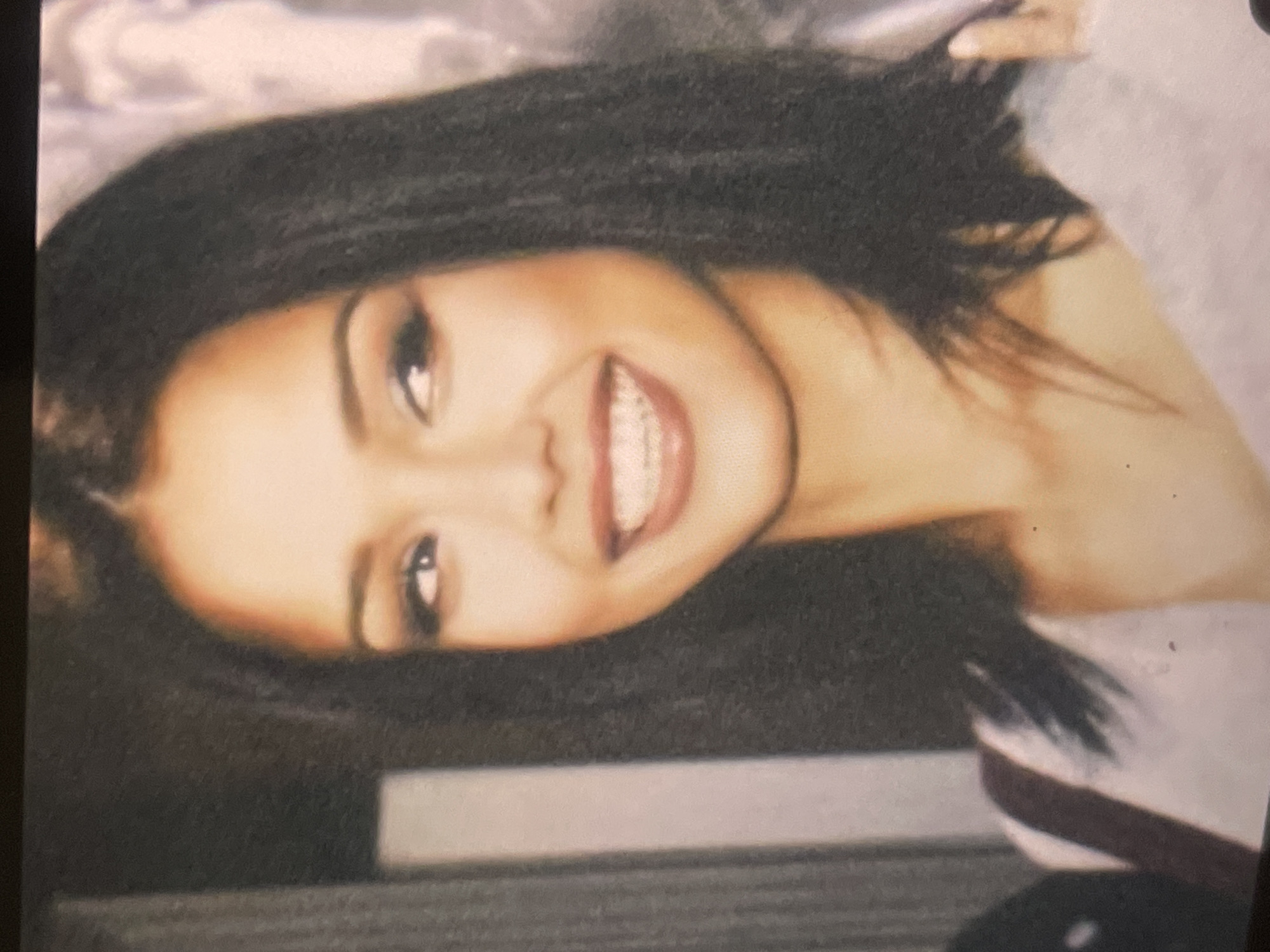A vibrant portrait captures a young woman with dark, long hair, smiling broadly and looking directly at the viewer. Her head is tilted, conveying a cheerful and friendly demeanor. She has fair skin, bright eyes with subtle makeup, and a wide, open-mouthed smile that reveals white teeth and full, reddish-pink lips.
Her dark hair appears straight or slightly wavy, framing her face and falling over her shoulders. She is wearing a light-colored top, possibly a sweatshirt or sweater, partially visible on her right side. The background is softly blurred with muted, light tones, suggesting an indoor setting with a simple backdrop. The overall image has a warm color cast and a slightly grainy quality, indicative of a scanned or photographed physical print. There is no visible text. The context for this image is Poltava, Ukraine.