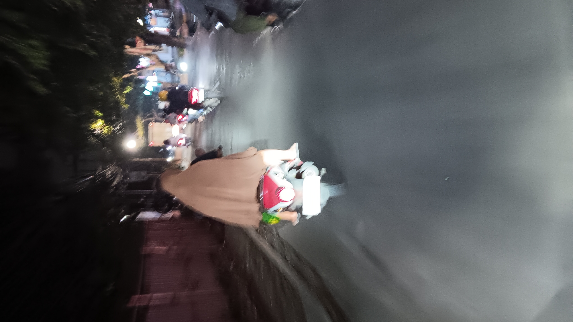 An outdoor, nighttime, medium shot shows several motorcycles traveling on a paved road in Depok, Indonesia. The image is blurry due to motion. In the foreground, a person wearing a brown headscarf and a green garment rides a motorcycle. The motorcycle is red and white with a white license plate. Several other motorcycles are visible in the background, with their headlights illuminating the road. Trees line the right side of the road, and a building with a pink fence is on the left. The sky is dark, indicating it is nighttime.