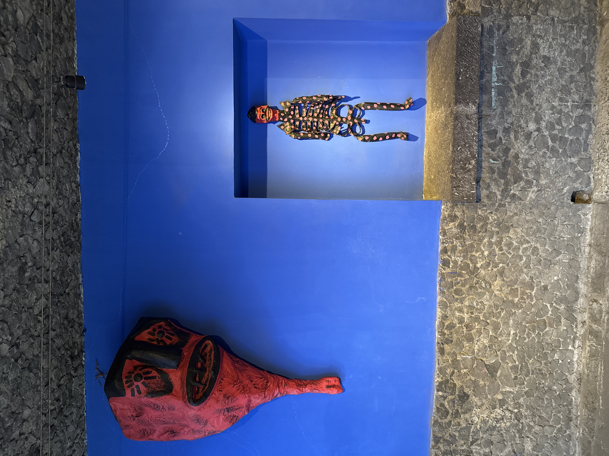 The image shows a section of a wall, presumably in a museum or gallery in Mexico City, Mexico. The wall is painted a vibrant blue color. Two art pieces are displayed on the wall. The first is a sculptural figure of a skeleton, painted in black with pink polka dots, lying within a recessed blue compartment. The second is a red and black mask, shaped like a foot. The background is a gray, rough stone wall. A thin cable runs down the stone wall. The art pieces are illuminated by the lighting in the space.