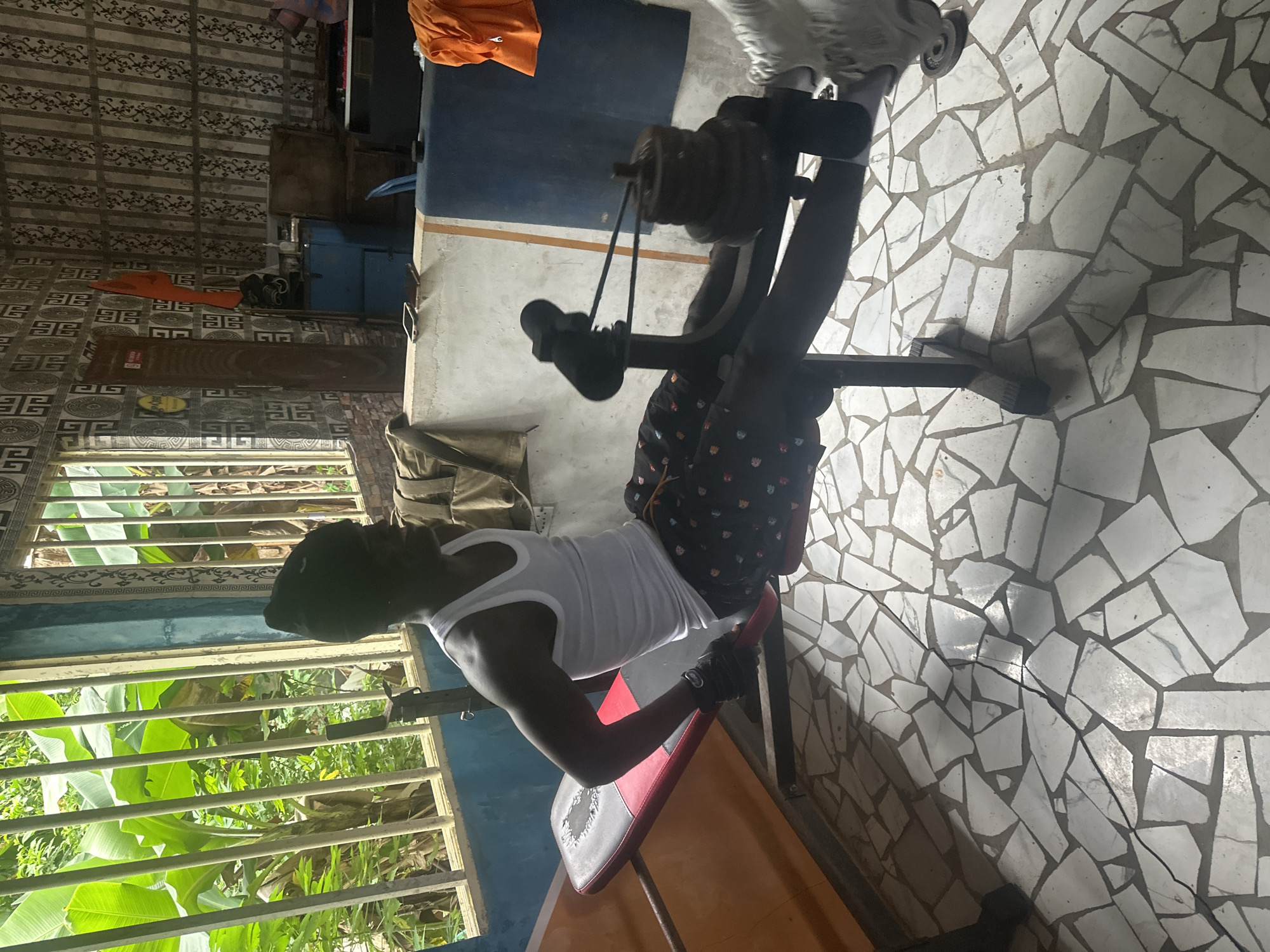 An unidentifiable male wearing a white tank top, black beanie, and patterned shorts is performing a seated leg extension exercise. He is positioned on a weight bench with a pulley system attached to weights. The exercise bench is situated on a floor tiled with irregular white and grey pieces. The scene appears to be an outdoor or semi-outdoor gym space. Behind the man, there is a window with vertical bars looking out onto lush green vegetation, suggesting a tropical environment. The background also shows a patterned wallpaper, a blue cabinet, and some orange fabric draped over a piece of furniture. The overall impression is of a rustic, improvised gym in a potentially humid climate, possibly in Badagry, Nigeria, given the natural elements and the style of the building.