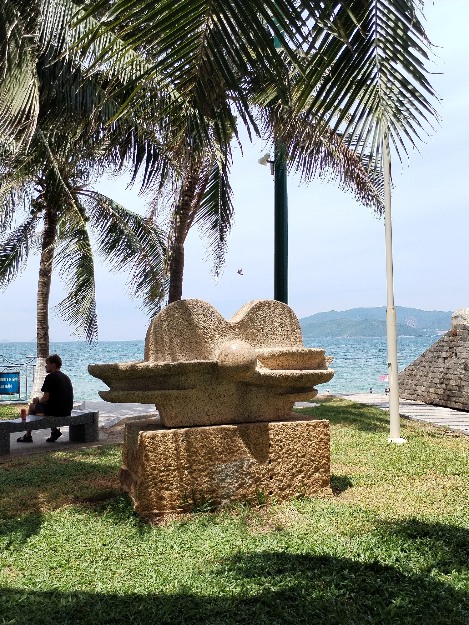 The image depicts a seaside scene in Nha Trang, Vietnam. In the foreground, a textured stone sculpture sits on a stone base amidst lush green grass. To the left, a man wearing a black t-shirt and shorts is seated on a stone bench, facing away from the camera. A red can is placed on the bench next to him. Behind the bench, a blue sign with text is visible. The middle ground features palm trees with their fronds casting shadows on the ground. Beyond the trees, a tranquil blue ocean stretches towards the horizon, where distant hills are faintly visible. A few people can be seen in the water, and a pink umbrella is set up on the beach. To the right, a textured stone wall borders a paved walkway. The sky is clear and bright, suggesting a sunny day.