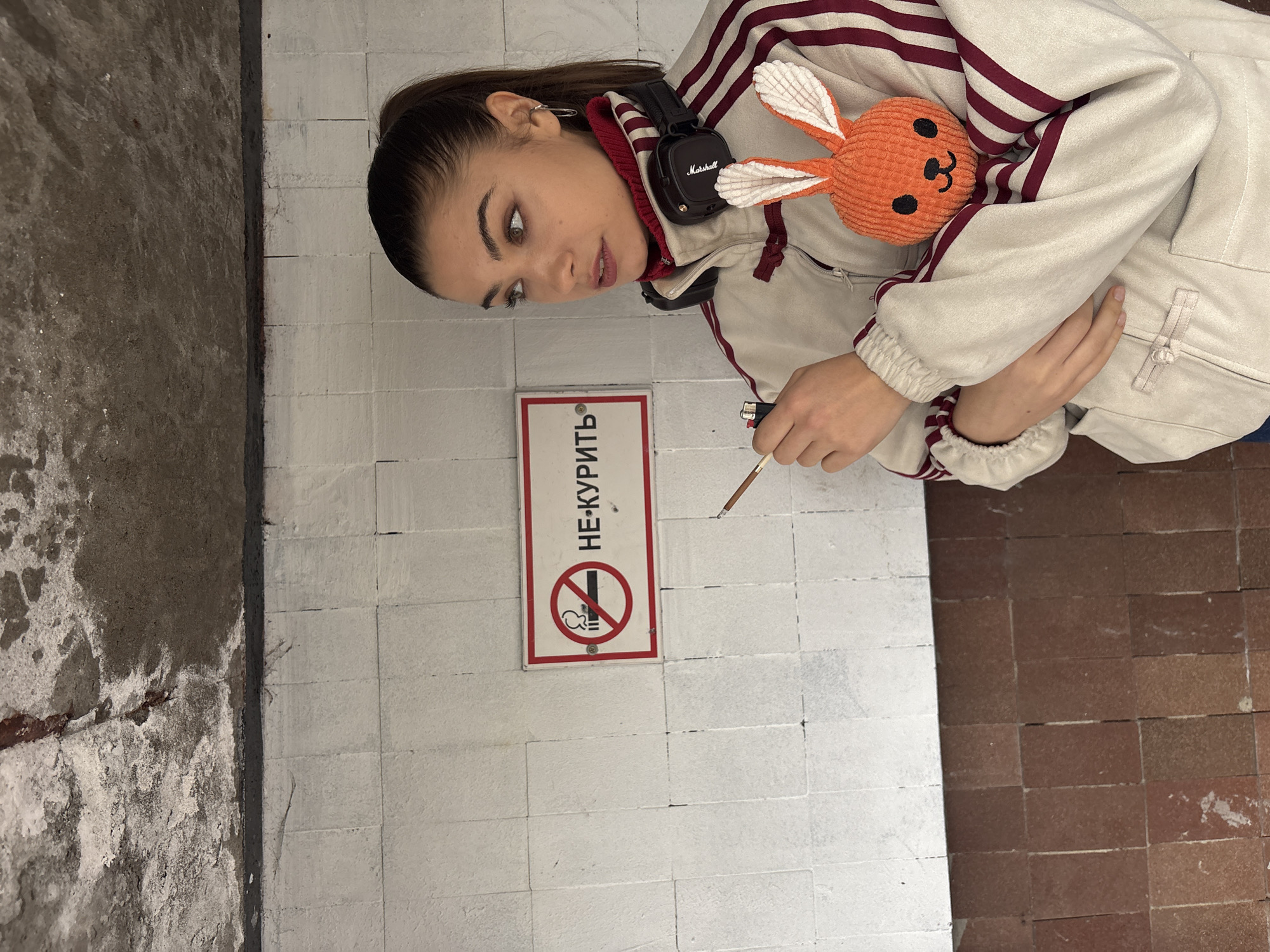 A young woman is standing next to a "No Smoking" sign, with the Cyrillic words "НЕ-КУРИТЬ" in Moscow, Russia. She is holding a lit cigarette and a lighter. She has headphones around her neck and is carrying an orange plush bunny. The background consists of white tiled walls and a reddish-brown tiled floor. A concrete wall is to the left.