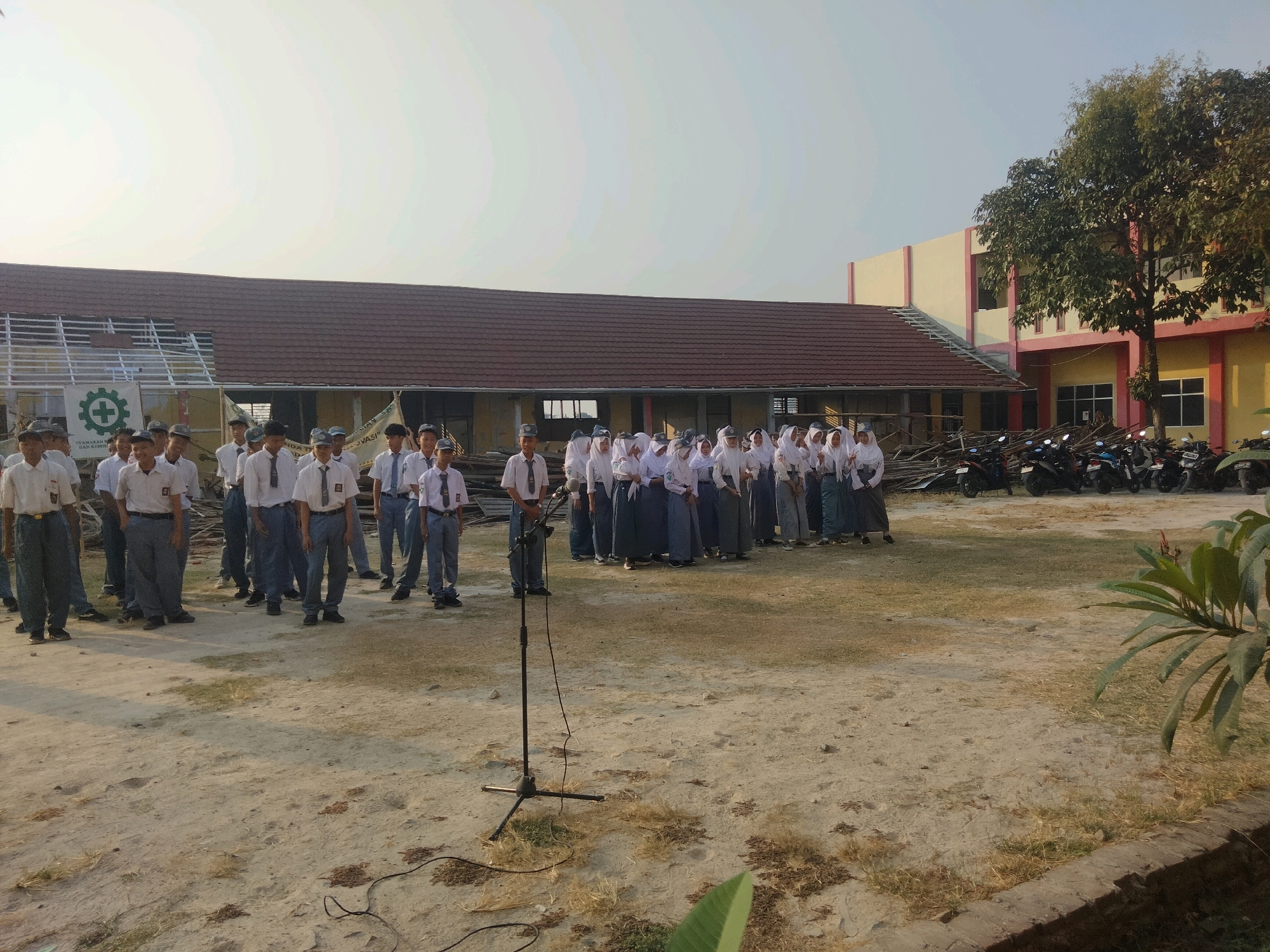In Suka Sari, Indonesia, a group of students in uniform stand in a dusty outdoor area in front of a building. The students, some wearing white shirts and ties with grey pants, and others wearing white shirts and grey skirts or hijabs, are arranged in rows. A microphone on a stand is positioned in the foreground. In the background, a building with a red tiled roof and yellow and pink accents is visible. Motorcycles are parked in front of the building. The ground is covered in dirt and dry grass. The sky is bright and clear.