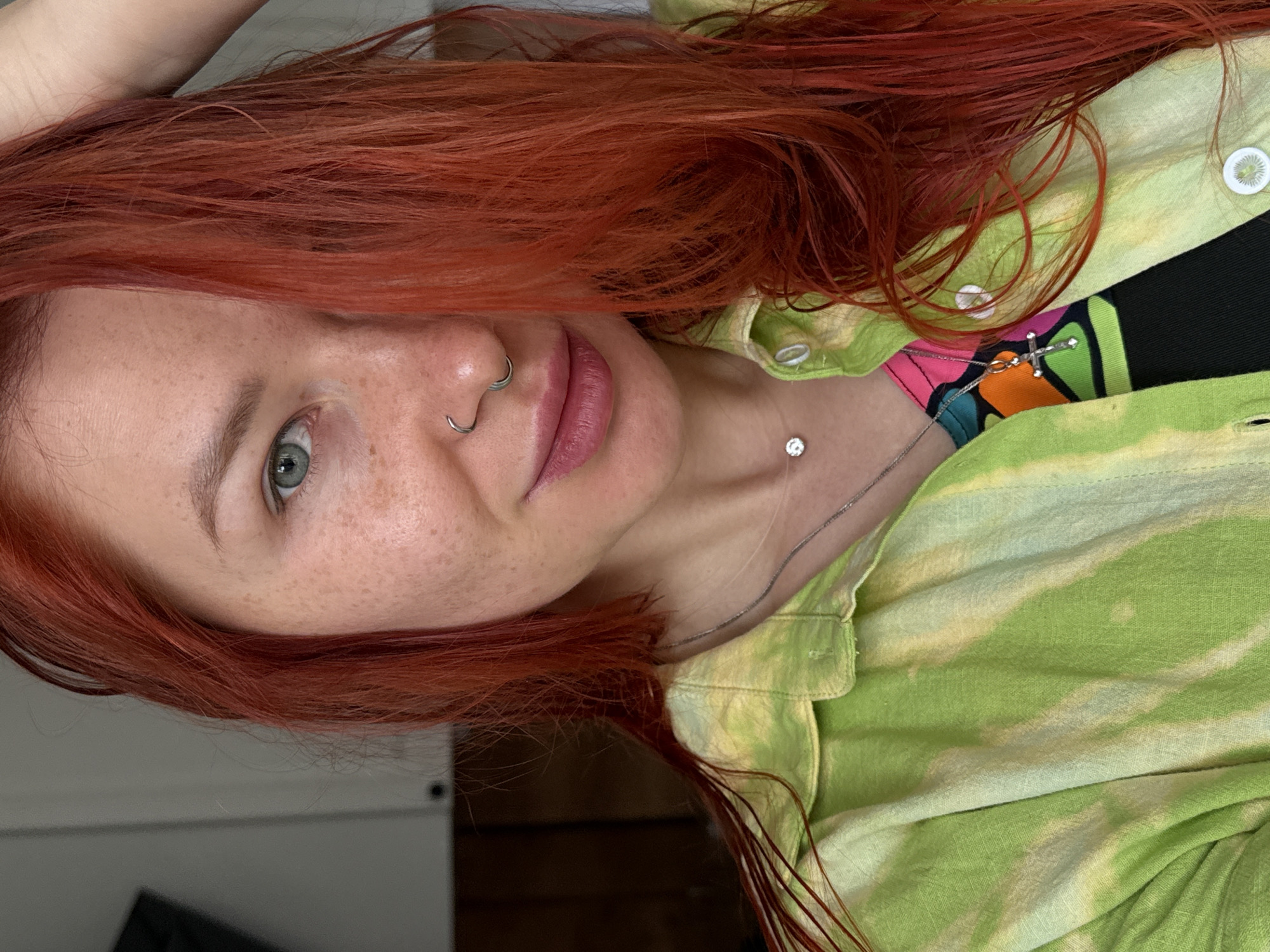 A young woman with bright red hair and fair skin poses for a close-up shot. She has freckles on her face, blue eyes, and two nose rings. She is wearing a green and yellow tie-dye shirt. A thin silver necklace with a small pendant is visible around her neck, and a colorful top is seen beneath the shirt. The background is blurred, suggesting an indoor setting. I don't see anything that indicates the location is Rostov-on-Don, Russia.