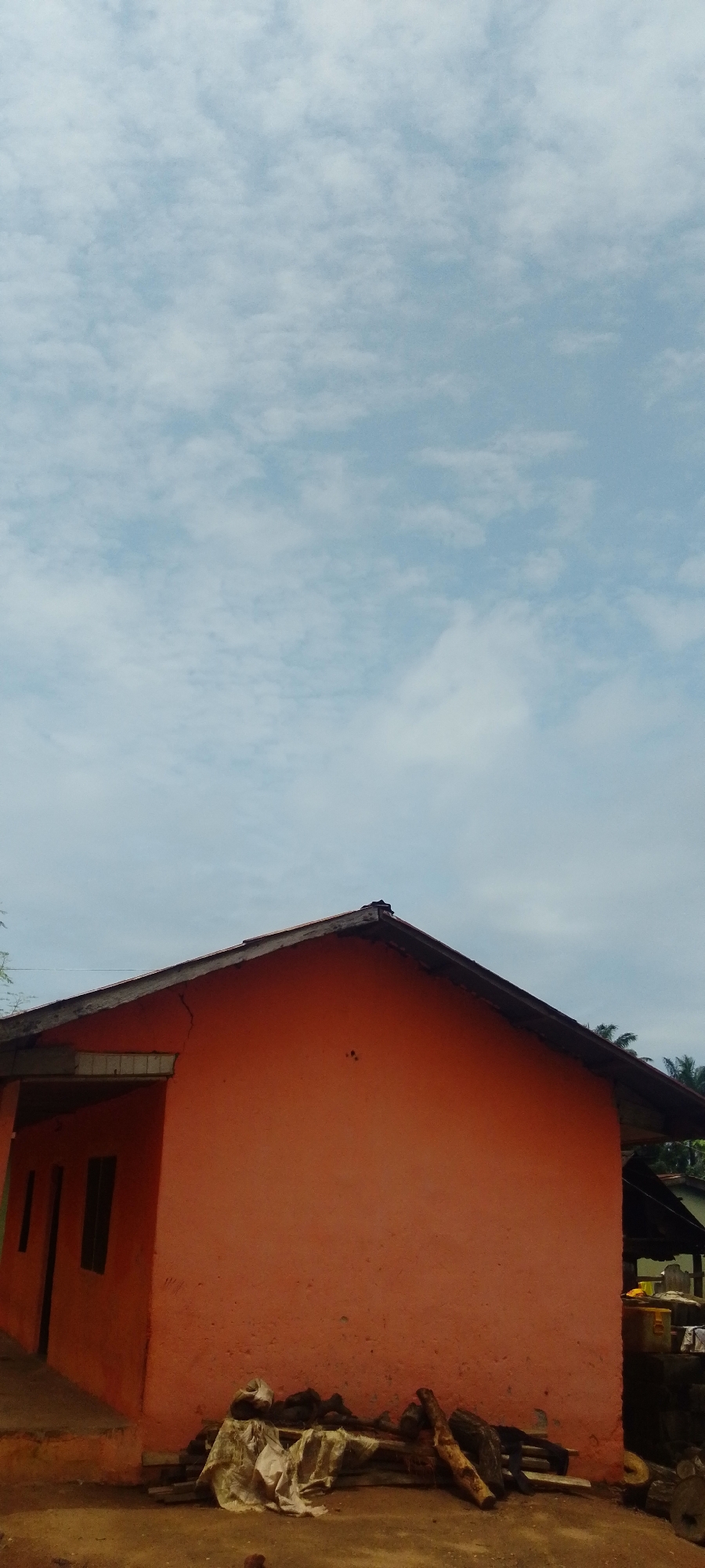 The image captures a side view of an unfinished or weathered orange building under a cloudy sky. The building has a simple, pitched roof with exposed wooden beams along the edge. On the left side of the building, there are two dark rectangular openings that appear to be windows or doors. The exterior wall of the building shows some cracks and a slightly rough texture.
In front of the building, the ground is uneven and dusty, with some scattered debris including pieces of wood, cloth, and possibly other building materials. Some of these materials are piled together, suggesting they might be stored or awaiting use. Further to the right, partially visible behind the main structure, is another darker roofed area or shed, with some yellow and white objects that could be containers. Palm trees are visible in the background, suggesting a tropical or sub-tropical environment.
The sky is filled with a layer of diffused white and grey clouds against a pale blue background, indicating an overcast or partly cloudy day. The lighting suggests it is daytime, possibly late morning or early afternoon, with diffused sunlight. There is no text visible in the image. The overall scene conveys a sense of rural simplicity, possibly in a developing area, given the unfinished look of the building and the surrounding conditions. There are no people visible in the image.