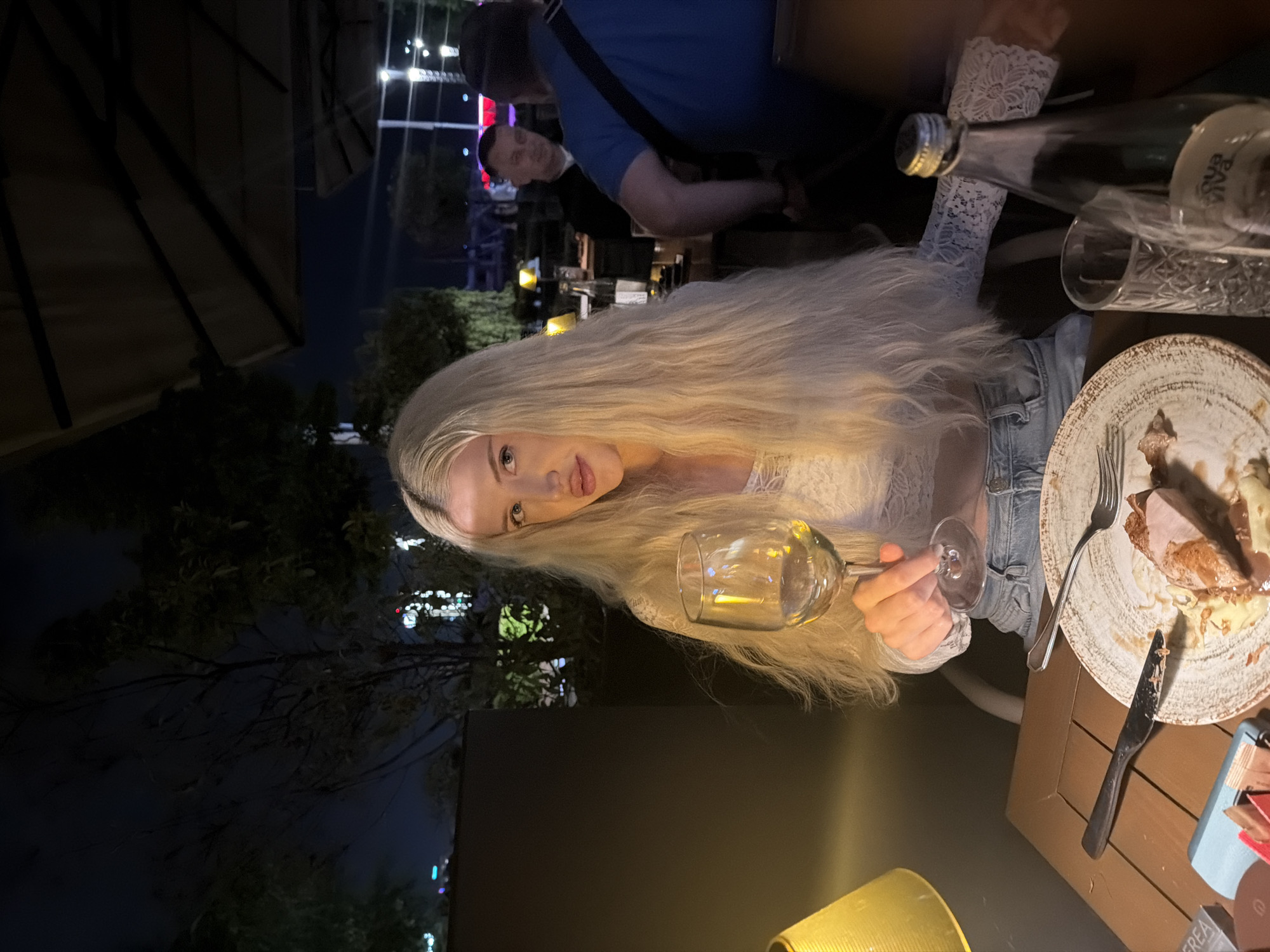 A woman with long, blonde hair is seated at an outdoor table in what appears to be a restaurant or bar, holding a wine glass filled with a light-colored liquid, presumably white wine. She is wearing a white lace top and a denim jacket. The table is set with a plate of food, a fork, and a knife. A bottle of water and a drinking glass are also visible on the table. Behind her, another person in a blue shirt is standing. A second male is slightly visible in the background. Trees and lights suggest that the location is outside. The background includes some streetlights or building lights, which would suggest a restaurant in an urban setting. The location could be Belgrade, Serbia.