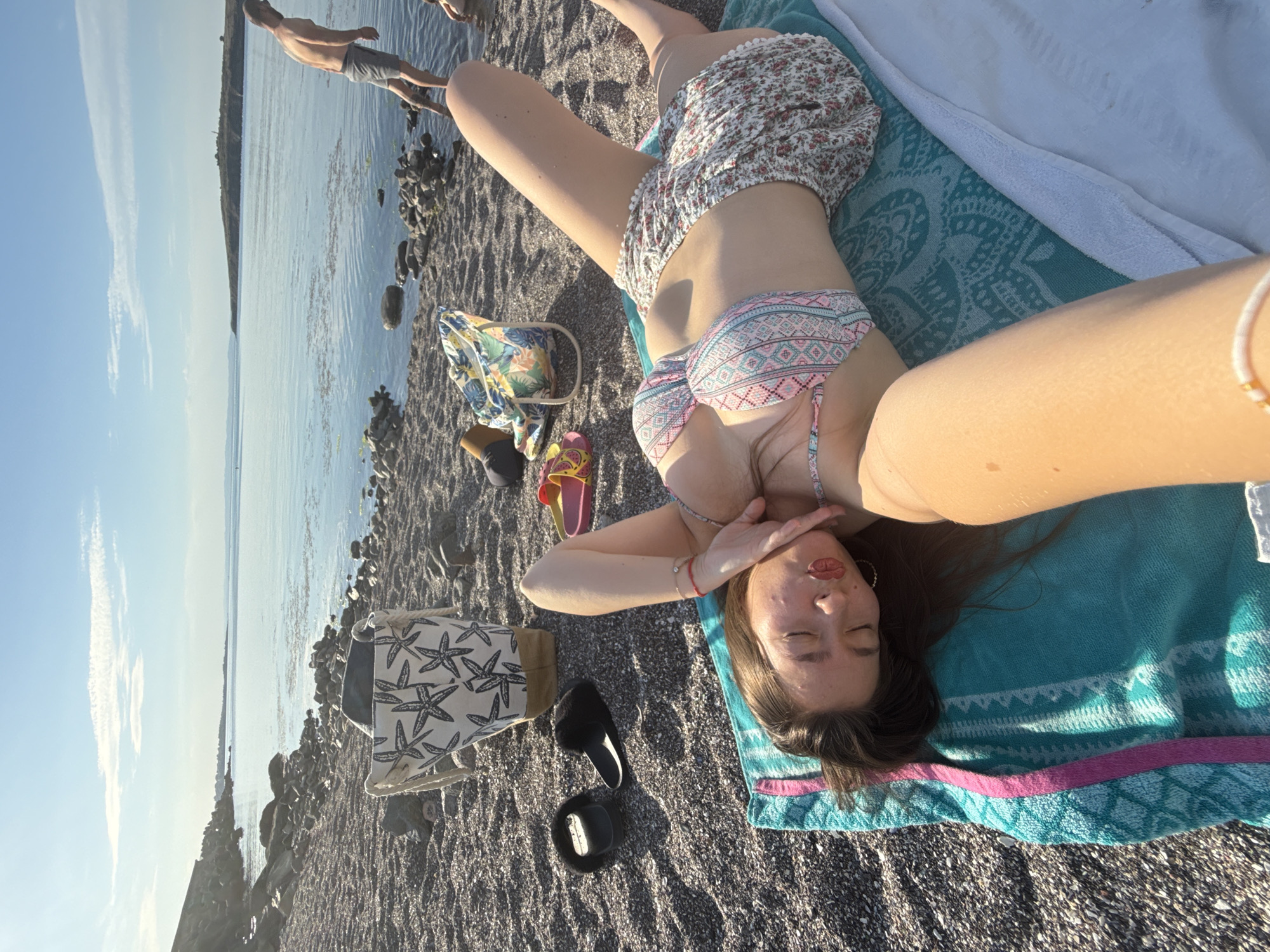 The image depicts a sunny day at a rocky beach in Sozopol, Bulgaria. In the foreground, a young woman lies on her back on a turquoise towel, wearing a patterned bikini and floral shorts. Her eyes are closed, and her hand is near her face, suggesting relaxation. To her left, there are black fuzzy slides and yellow and pink flip-flops. Behind her, a patterned beach bag with starfish is visible. The beach is covered in dark pebbles and rocks. The sea stretches out in the background, with a faint outline of land in the distance. A man in gray shorts stands in the water on the right side of the image, his back to the camera. The sky is clear blue with a few wispy clouds.