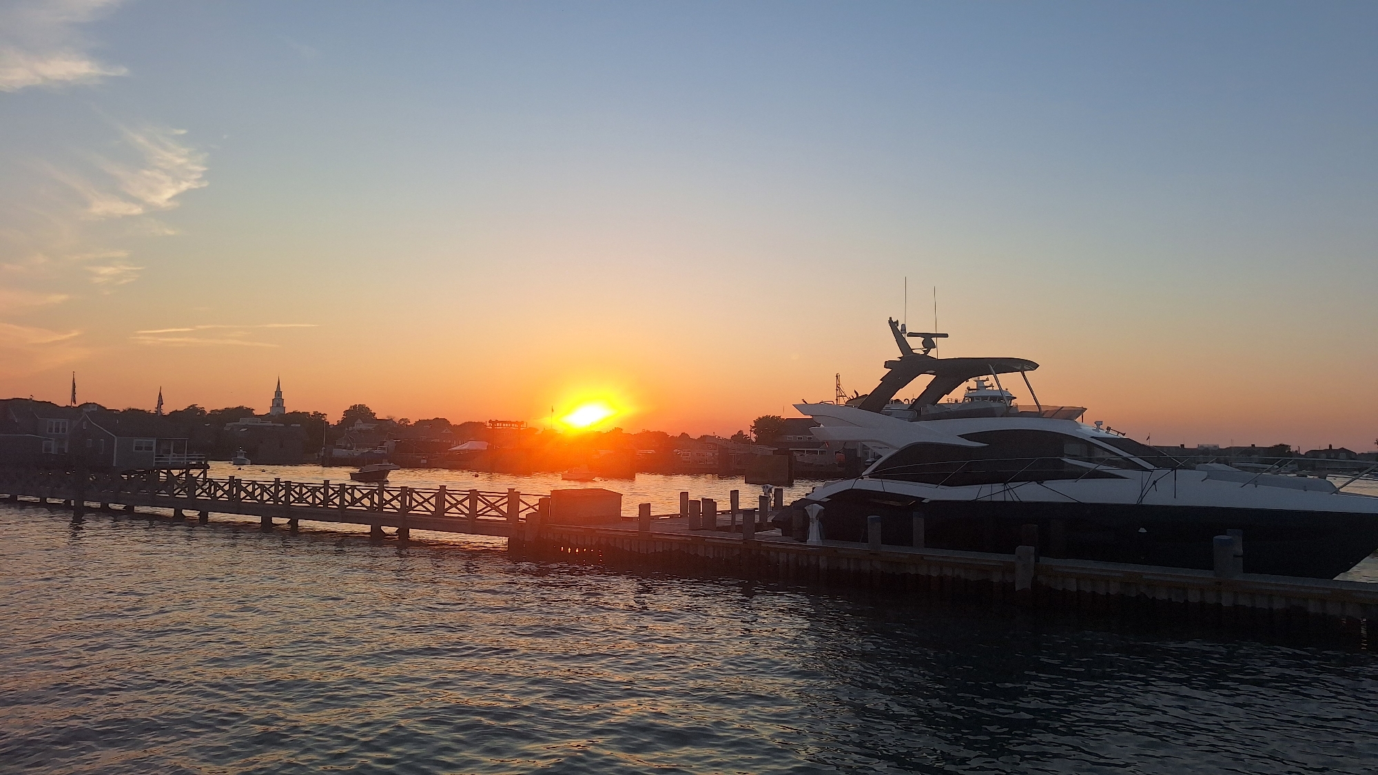 At sunset in Nantucket, Massachusetts, a large, modern yacht is docked at a pier. The yacht is dark blue and white, with a sleek design. It has a tall mast with antennas. The pier is made of wood and has railings. The water is calm and reflects the colors of the sunset. In the background, there are several other boats, buildings, and a church steeple. The sky is a gradient of blue and orange, with a few wispy clouds. The scene is peaceful and serene.