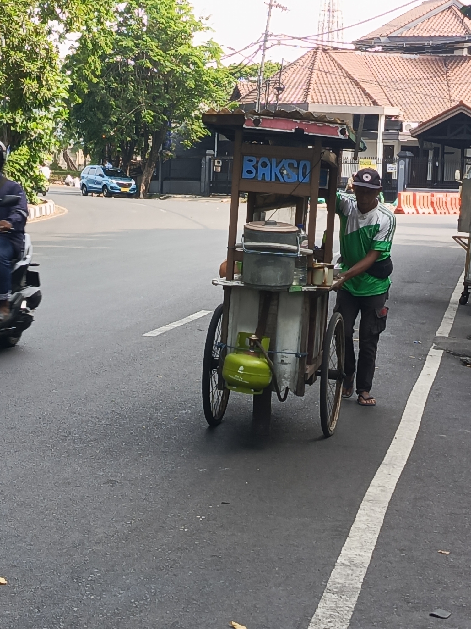 An elderly man with grey hair and a green and white striped t-shirt pushes a bakso cart down a street in Surabaya, Indonesia. The cart is wooden and has a red and white awning with the word "BAKSO" painted in blue letters. A green propane tank is strapped to the side of the cart, and various metal containers and bottles are visible on shelves. The man is wearing dark pants and sandals. In the background, a blue car is driving on the road, and trees and buildings line the street. The weather appears to be sunny and bright.
