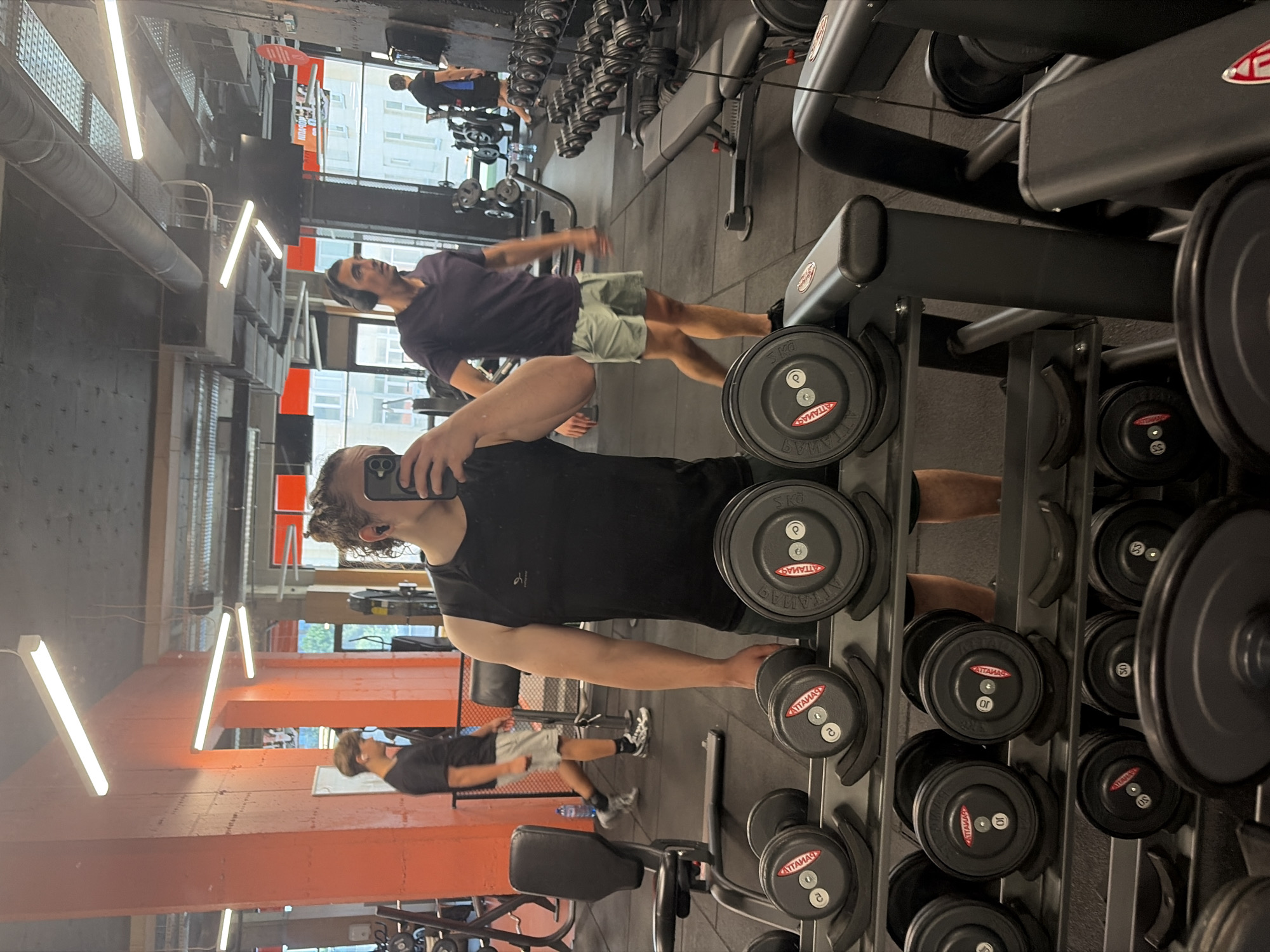 A gym interior in Plovdiv, Bulgaria is observed. Several individuals engage in exercise activities. A man is taking a photo of himself in the foreground. Dumbbells and weightlifting machines are visible. The lighting appears artificial, and the gym has a modern design.