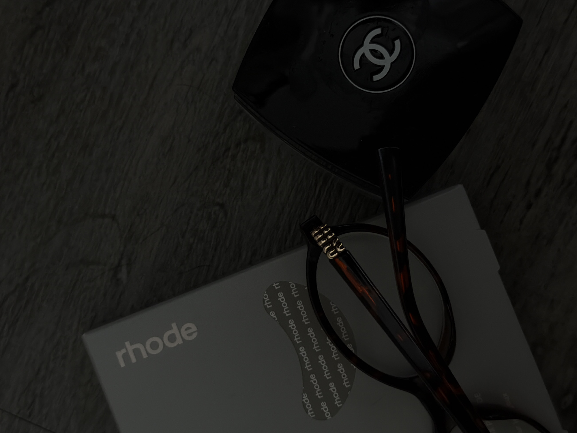 The image is a dimly lit, close-up still life shot. In the upper right corner is a black, glossy Chanel compact with its iconic interlocking "C" logo in white. Below and slightly to the left of the compact is a gray box labeled "rhode". It has the word "rhode" written largely at the left with the rest of the copy in smaller, repeated iterations of the word "rhode". Resting on top of the "rhode" box are a pair of tortoise shell eyeglasses. The background appears to be a dark, textured surface. The scene is indoors with a focus on personal luxury and beauty items. The location is not evident in the image; it is possible that it is in Los Angeles, United States of America, as prompted.