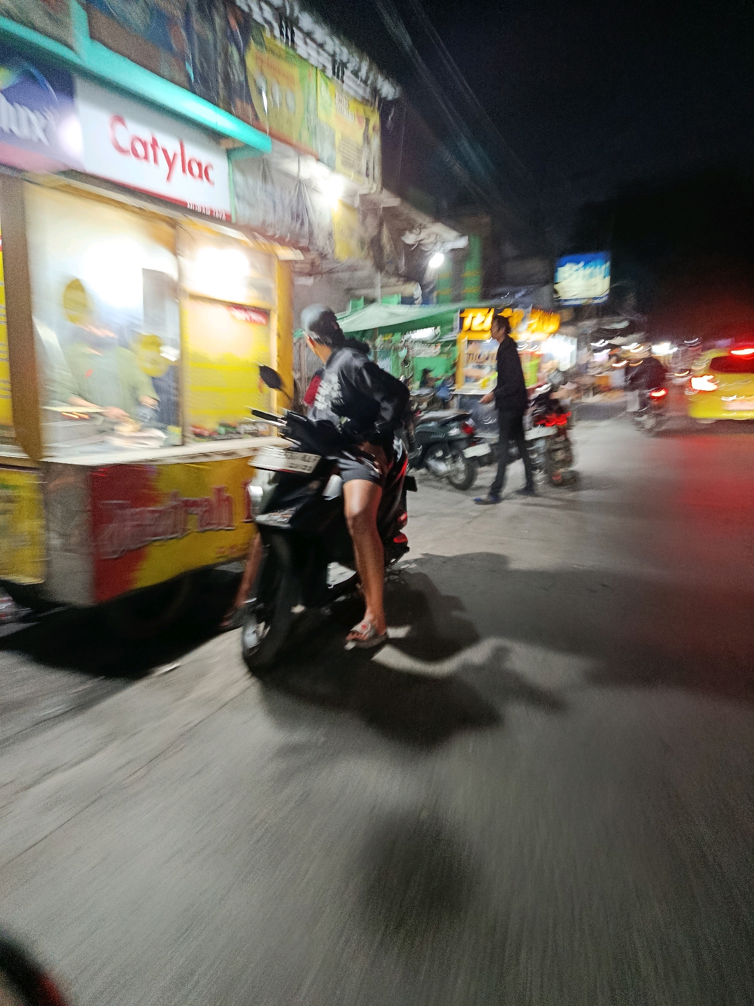 The image captures a street scene at night, likely in Bekasi, Indonesia, with several individuals and vehicles. A person is seated on a dark-colored motorcycle in the foreground, facing away from the camera. They are wearing a dark jacket and shorts, with one leg extended and resting on the ground. The motorcycle's license plate is partially visible.
To the left, a food stall is illuminated, with signage that includes the word "Catylac" in red lettering. Inside the stall, at least two people are visible, one appearing to be preparing food. The stall has a yellow and red facade with writing on it.
In the background, other motorcycles and vehicles are present, along with brightly lit signs and streetlights, indicating an urban environment. Motion blur is evident throughout the image, suggesting movement and a low-light photographic setting. The overall impression is of a bustling nighttime street in a commercial area.