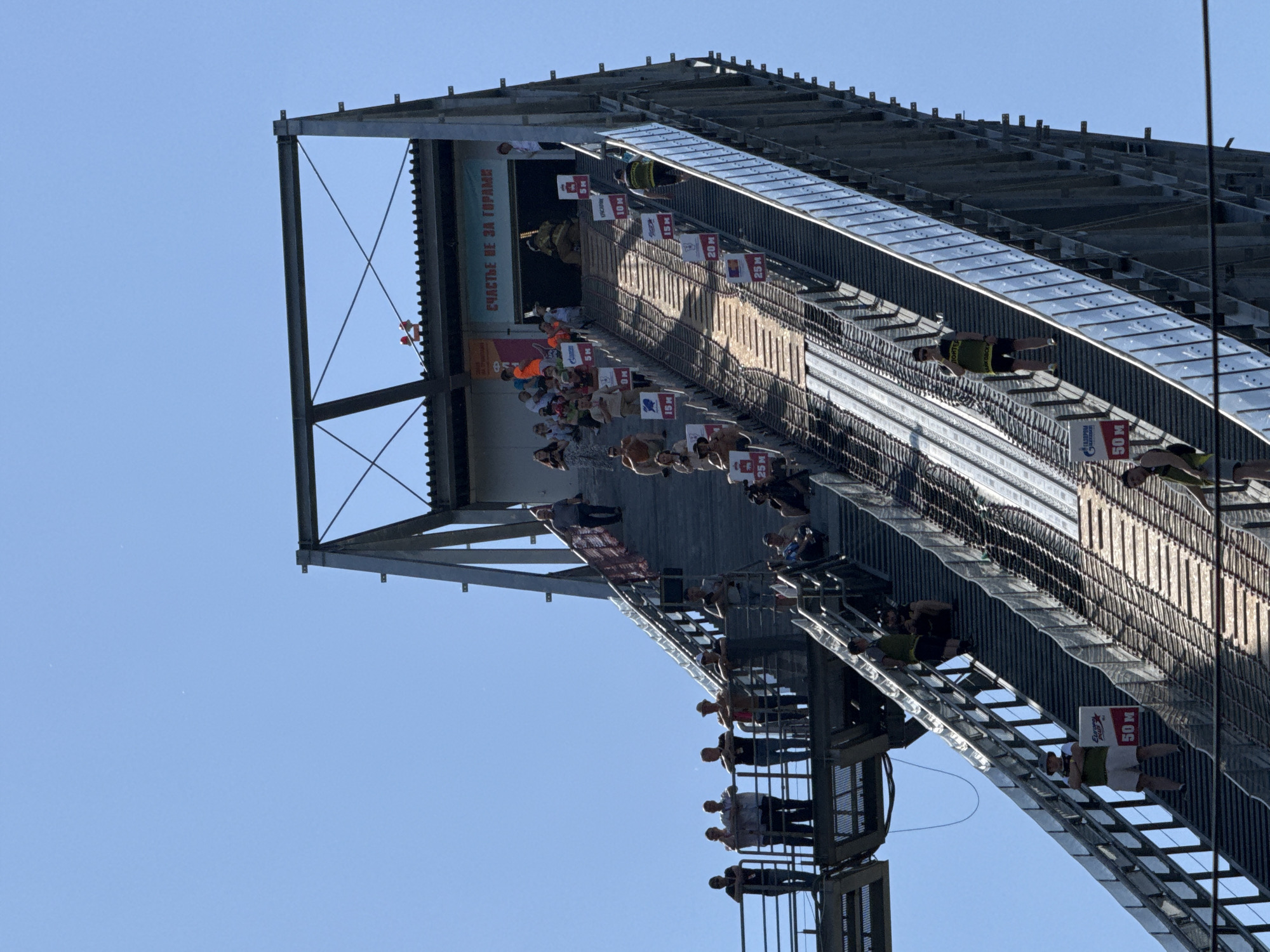 The image captures a high diving platform under a bright blue sky. The platform appears to be constructed of metal with multiple levels. The levels are marked with distances, with one clearly labeled "50 M" on a sign. Multiple people are seen on the platform at various levels, seemingly divers preparing to jump. Some spectators are also visible. A banner with text in a language other than English is affixed to the platform. The city is labeled as Unknown, Unknown.