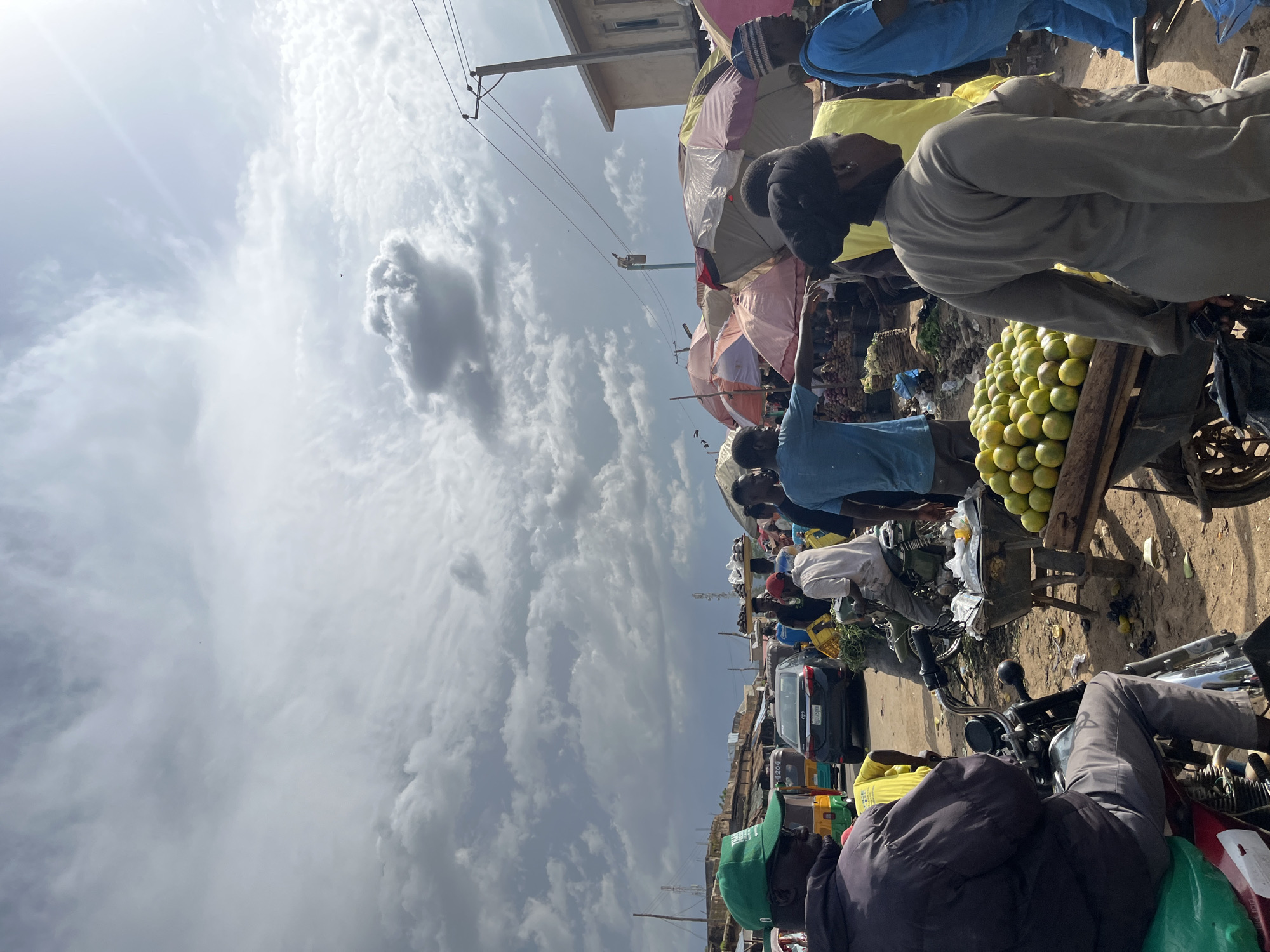The image depicts an outdoor market scene in Rafin Par, Nigeria. The sky is filled with dramatic, grey clouds, suggesting an overcast day, with some patches of bright light. In the foreground, a man wearing a green cap and a dark jacket is seated on a motorcycle. To his right, a cart filled with oranges is visible, with a woman in a grey outfit standing nearby. In the background, numerous people are milling about, some under colorful umbrellas and awnings. Several vehicles, including motorcycles and a car, are present, indicating a busy thoroughfare or intersection within the market area. Stalls with goods are set up, and there are indications of various vendors and shoppers engaged in activity. The overall impression is of a lively, bustling marketplace.
