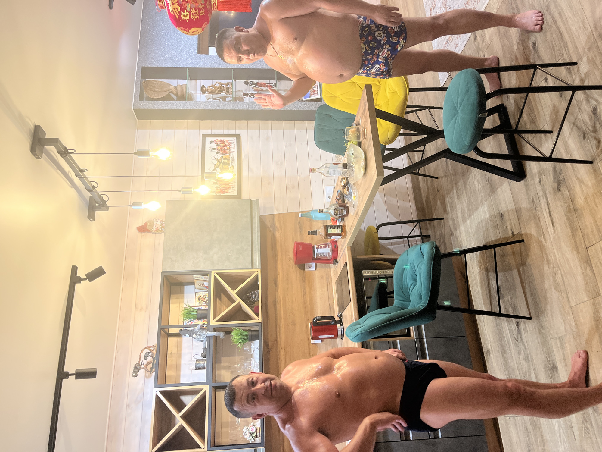 Two shirtless men wearing underwear stand in a kitchen in Новая Чернушка, Russia. The kitchen has wooden floors and cabinets, a dining table with colorful chairs, and various kitchen appliances. The room is lit by overhead lights.