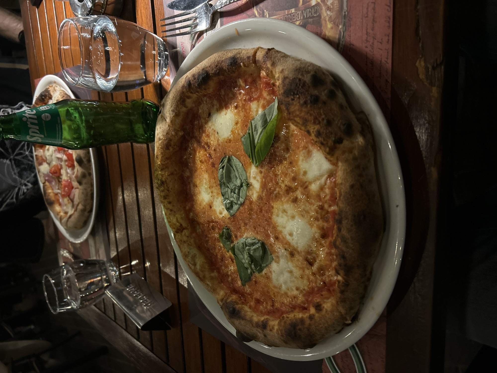 The image depicts a table setting, likely in a restaurant in Plovdiv, Bulgaria. A pizza with tomato sauce, mozzarella, and basil leaves rests on a white plate. Next to it are a green Sprite bottle, a glass, and cutlery. Another pizza is visible in the background on a separate plate. The table itself is made of wood and appears to be outdoors or in a dimly lit area.