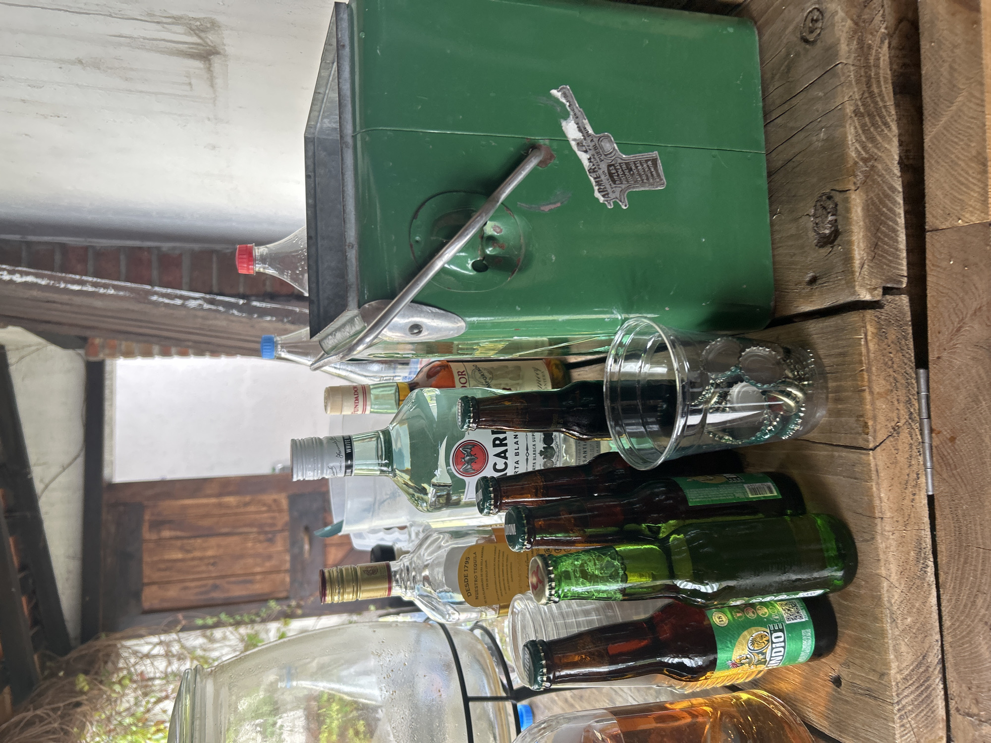 A still life photograph captures a cluttered bar scene, composed of various alcoholic beverages and containers. Stacked on a weathered wooden surface are bottles of tequila, rum (Bacardi Carta Blanca), beer (Indio), and other unidentified liquors. A clear plastic cup overflows with bottle caps. Atop the alcohol is a green ice chest, adorned with a sticker referencing rifles, pistols, and explosives, with the location indicated as Puebla, Mexico. The background reveals a partially visible exterior, including portions of a building's architecture and vegetation.