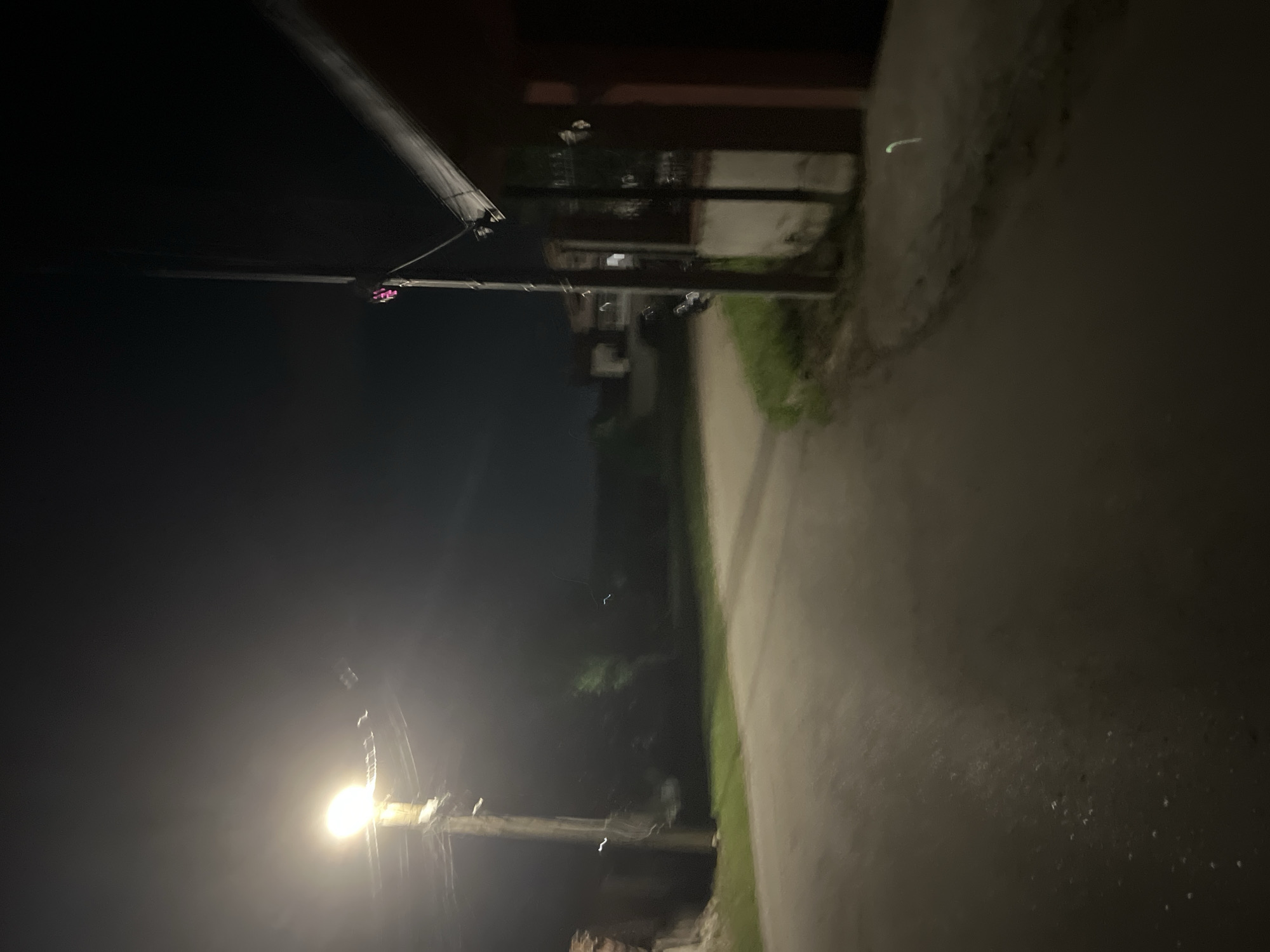 The image depicts a nighttime scene on a paved road in the city of Unknown, Unknown. A prominent, intensely bright spherical streetlamp, mounted on a wooden utility pole, illuminates the left side of the foreground and a section of the road. Multiple electrical wires extend from this utility pole. The road itself is dark asphalt, appearing somewhat reflective or wet, with scattered small, bright specks on its surface. It curves gently to the right as it recedes into the dark background.
On the right side of the frame, a dark, reddish-brown building with a sloped roofline is visible, partially obscured by a second, darker utility pole. This second pole also supports electrical wiring and features a distinct small, pink or magenta light source attached to its upper section. Vertical structural elements are discernible on the building's facade, along with a reflective or windowed area. Patches of unkempt grass line both sides of the road near the foreground structures.
The background is largely undifferentiated darkness, with faint, blurred outlines suggesting distant structures or foliage. Several thin, wavy, white light trails are present in the air, particularly near the illuminated streetlamp and further down the road on the left. A single, distinct, curved green light trail is visible on the ground near the lower right corner of the frame. No individuals, animals, or explicit actions are discernible within the scene.