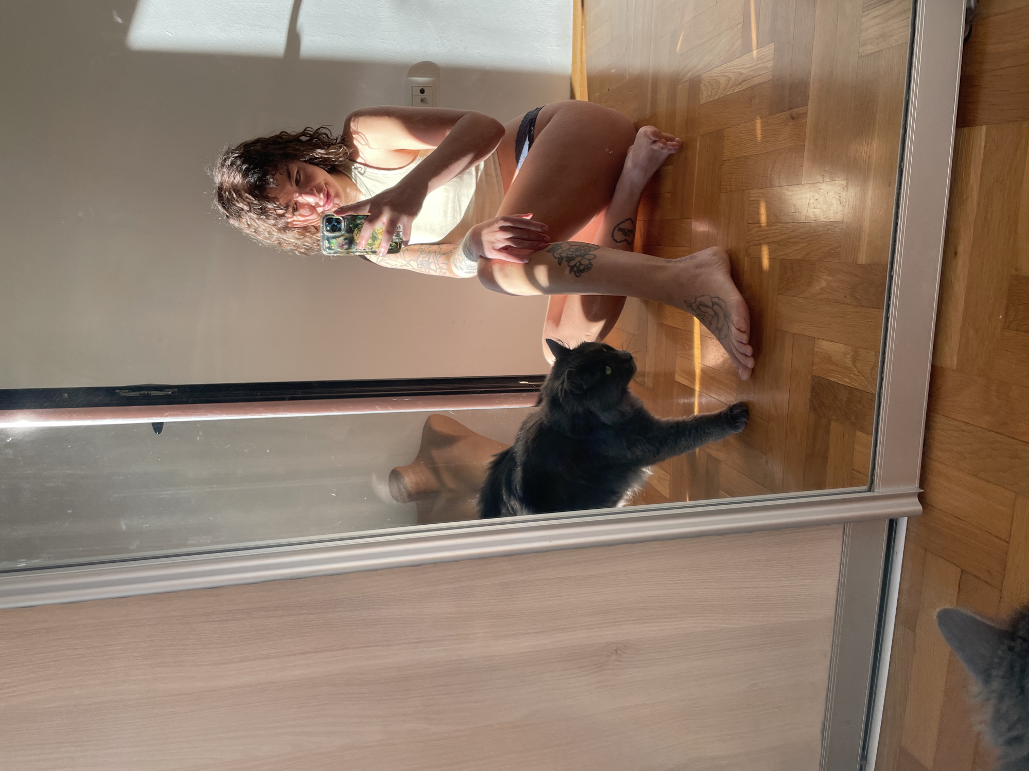 The image depicts a woman taking a mirror selfie in an indoor setting, presumably in Boreti, Montenegro. The woman, with curly brown hair, is reflected in a large, full-length mirror. She is in a crouching or kneeling pose on a light-colored wooden parquet floor, wearing a light tank top and dark underwear. Her arms and legs are adorned with several tattoos, including a prominent floral design on her left calf and visible patterns on her right arm and shoulder. She holds a smartphone with a distinctively patterned case in her right hand, capturing her reflection.
Bright natural sunlight floods the room, casting long, sharp shadows across the white wall and the wooden floor, indicating it is daytime, possibly morning or late afternoon. An electrical outlet is visible on the white wall in the background. In the lower part of the mirror's reflection, a dark gray or black cat is seen sitting or lying down with one paw extended, seemingly looking towards the woman or the phone. Another cat's ear and part of its head are visible at the very bottom right, just outside the mirror's frame. The woman displays a relaxed and confident expression with a slight smile. The room features light wooden flooring and plain white walls, suggesting a cozy and well-lit domestic environment. No discernible text is visible in the image.