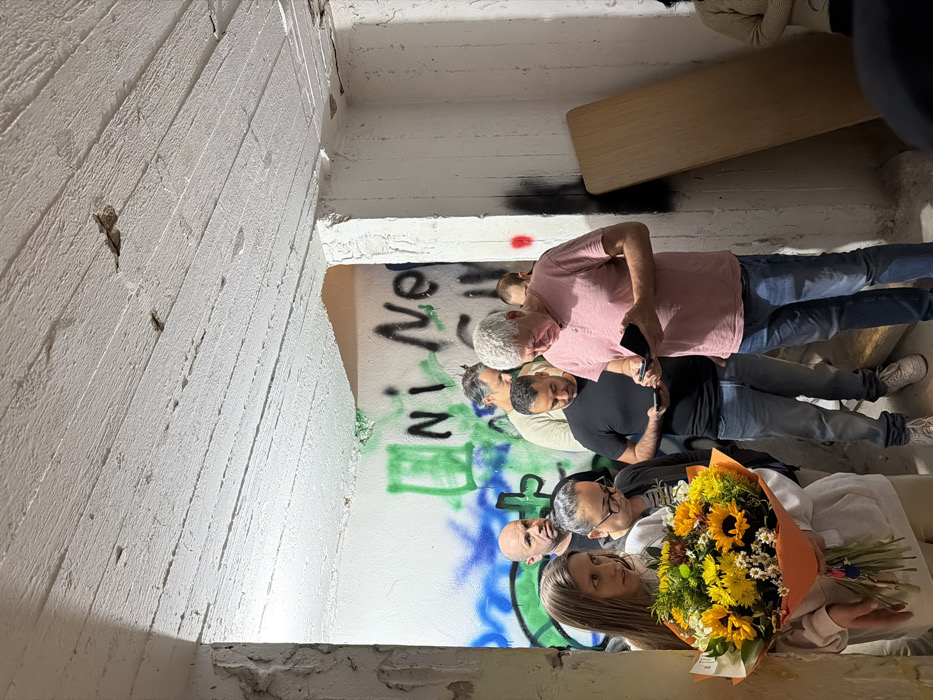 The image depicts an indoor gathering of approximately ten individuals within a stark, utilitarian space in Tel-Aviv, Israel, likely a basement or an unfinished concrete structure. The environment features a rough, white-painted concrete ceiling and walls, with exposed aggregate and prominent graffiti in green, black, and blue paint, including partial text like "Ni Ne." A doorway or passage is visible in the background. On the right, a light-colored wooden panel leans vertically against a wall.
In the foreground, a woman with long light brown hair, wearing a light-colored hoodie, stands on the left, holding a large bouquet of sunflowers and other yellow and orange flowers wrapped in orange paper, along with a set of keys. Her gaze is directed towards the right. Behind her, another woman with glasses is looking downwards. In the center foreground, an older man with white hair, dressed in a pink short-sleeved shirt and blue jeans, is focused on a dark object, possibly a wallet or phone, in his hands. To his left, a man with dark hair and a beard, wearing a black t-shirt and blue jeans, is engrossed in his smartphone. Other adult male figures are partially visible in the mid-ground and background, including a bald man in a dark t-shirt. Most individuals are standing, and several are engaged with personal electronic devices or objects in their hands. No direct interactions are depicted among the individuals.