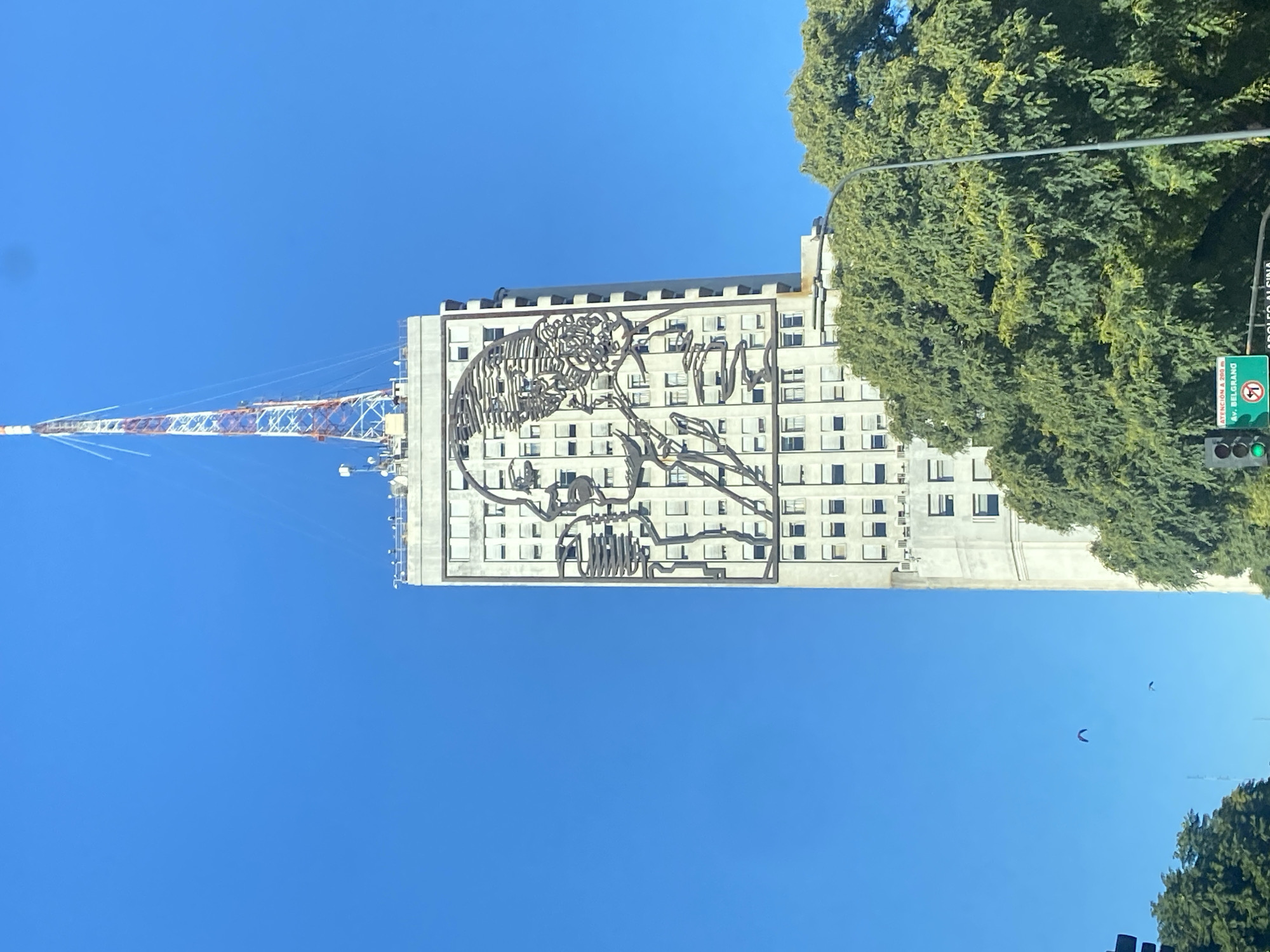 The image shows the Ministry of Social Development building in Buenos Aires, Argentina, located on Avenida 9 de Julio. The building has a metal sculpture of Eva Perón on its facade. A tall communication tower stands adjacent to the building. The sky is clear and blue. A tree is on the right side of the building. A traffic light and street sign are also on the right.