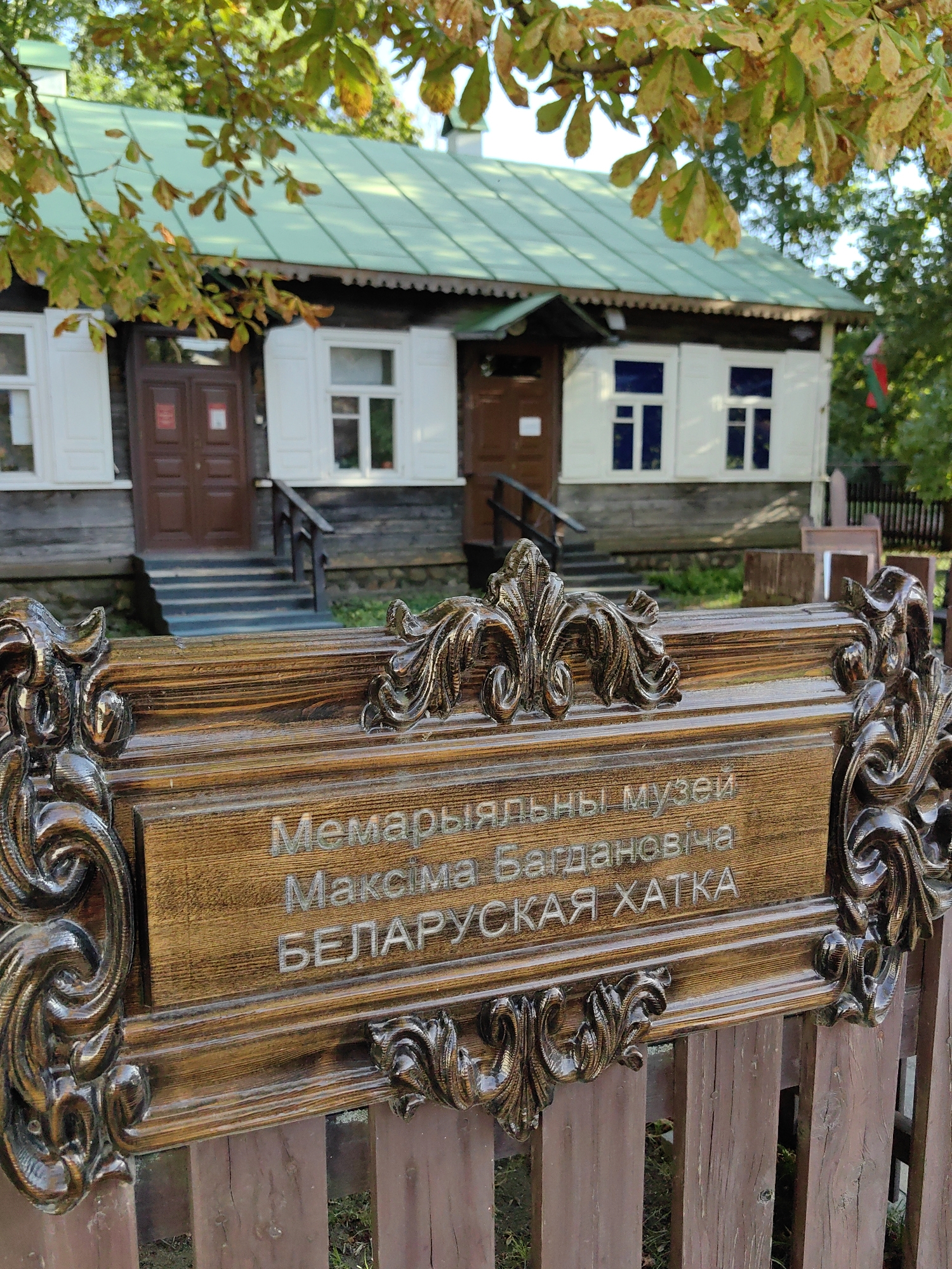 The image shows a wooden sign with an ornate frame in front of a traditional wooden house with a green roof. The sign is inscribed with text in Cyrillic. The text reads "МЕМАРИЯЛЬНЫ МУЗЕЙ МАКСІМА БАГДАНОВІЧА БЕЛАРУСКАЯ ХАТКА". This translates to "Memorial Museum of Maksim Bahdanovich "Belarusian Hut"". Maksim Bahdanovich was a famous Belarusian poet, and this appears to be a memorial museum dedicated to him, possibly his childhood home or a recreation of it. The house has white shutters on the windows and a wooden porch with stairs. There are trees with autumn leaves in the foreground and background. The overall scene suggests a cultural or historical site. It is located in Minsk, Belarus, at the Maksim Bahdanovich Literary Museum.