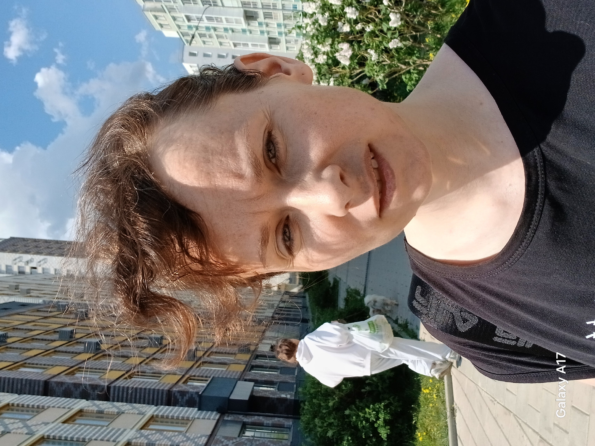 A woman with short brown hair is posing for a selfie on a sunny day. She is wearing a black shirt. In the background, there is a modern building with a distinctive yellow and grey facade. A person wearing white clothes and a white bag is walking with a white dog on a paved path. Greenery and flowers are also visible. The photo was taken with a Galaxy A17 phone. Based on the architectural style of the building in the background, it is highly likely that this photo was taken in the city of Yekaterinburg, Russia.