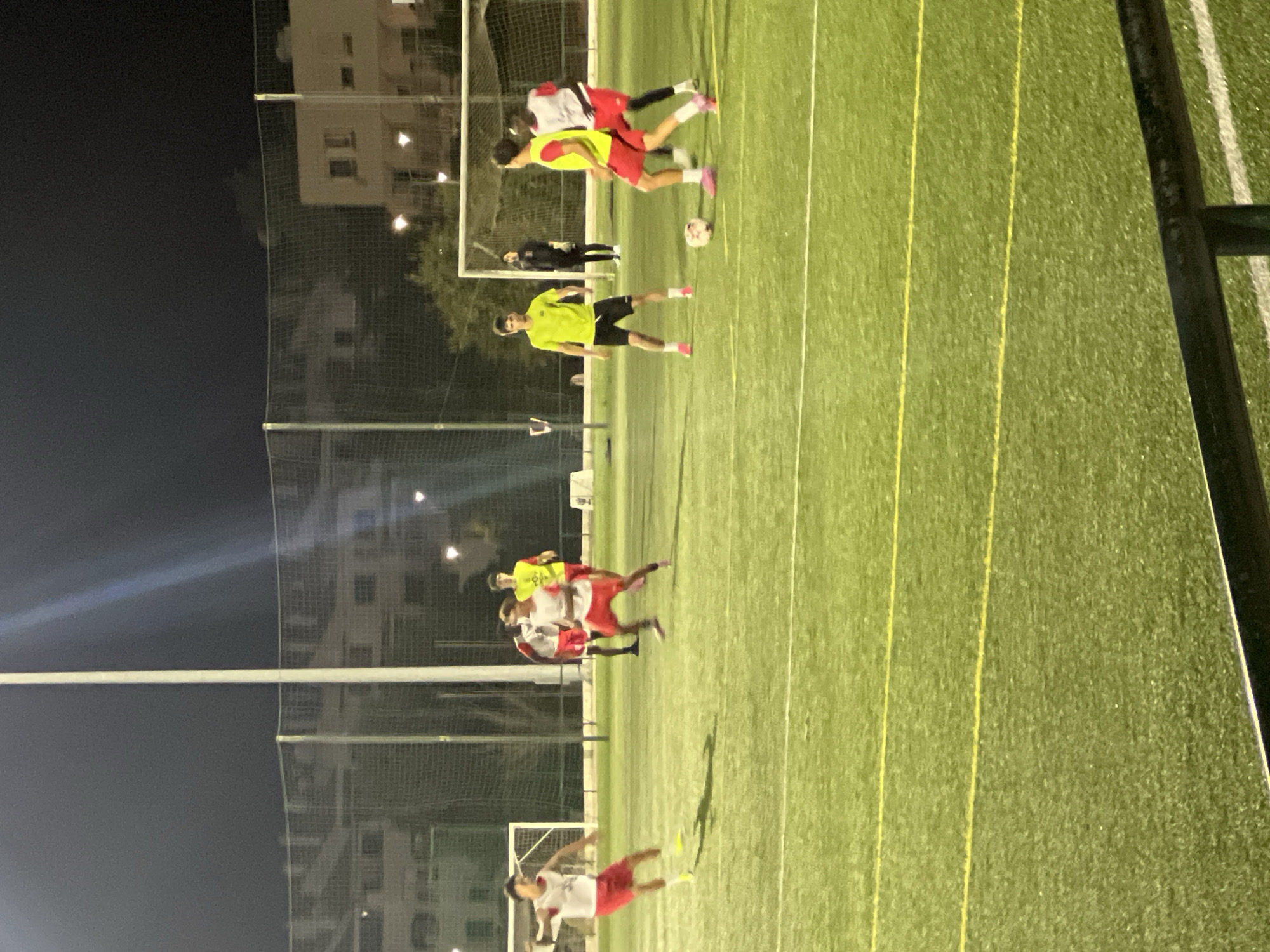 The image depicts a nighttime soccer game on an artificial turf field. Several individuals are actively engaged in play, wearing team uniforms. Two players in red shorts and white vests are on the left side of the frame, one in motion, the other further back. In the center, a player in a bright yellow shirt and black shorts stands near a soccer ball. Behind him, two players in red shorts and white vests are visible, one with a yellow vest over his shirt. Another player in a yellow vest and red shorts is in the foreground, also near the ball. In the background, a person in black clothing appears to be a goalkeeper. The field is illuminated by overhead lights, casting shadows on the turf. Beyond the field, a sports netting encloses the area, with buildings and trees visible in the distance under a dark sky. The location is identified as Albufeira, Portugal.
