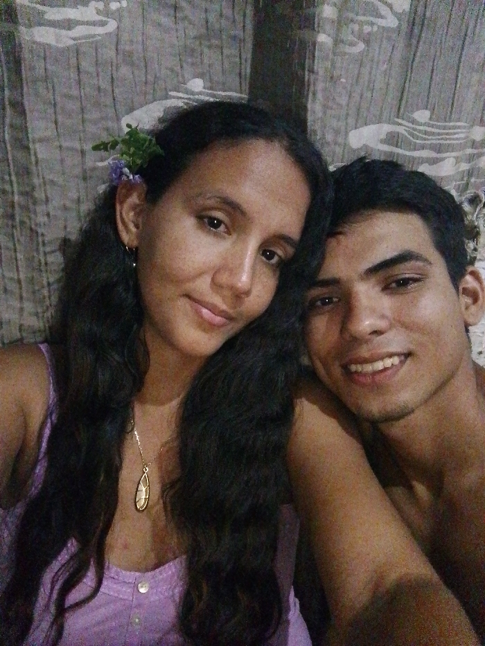 An indoor portrait depicts two young adults, a female and a male, in the city of Juan Griego, Venezuela. The female, positioned on the left, has long, dark, wavy hair, a dark complexion with visible freckles, and a light purple sleeveless top with buttons. Her left ear bears a small hoop earring, and a cluster of green leaves and faint purple flowers is arranged in her hair above her right ear. A gold-colored necklace with a tear-drop shaped pendant is visible around her neck. She looks towards the viewer with a slight, closed-lip smile. The male, positioned on the right and slightly behind, leans towards the female. He has short, dark hair and a dark complexion, and is shirtless or wearing an unidentifiable dark garment. He smiles, showing teeth, and also looks directly at the viewer. His right arm is partially visible on the female's left shoulder area. The background consists of a patterned fabric or curtain with abstract light-colored designs against a darker, mottled gray or brown surface. The lighting is dim.