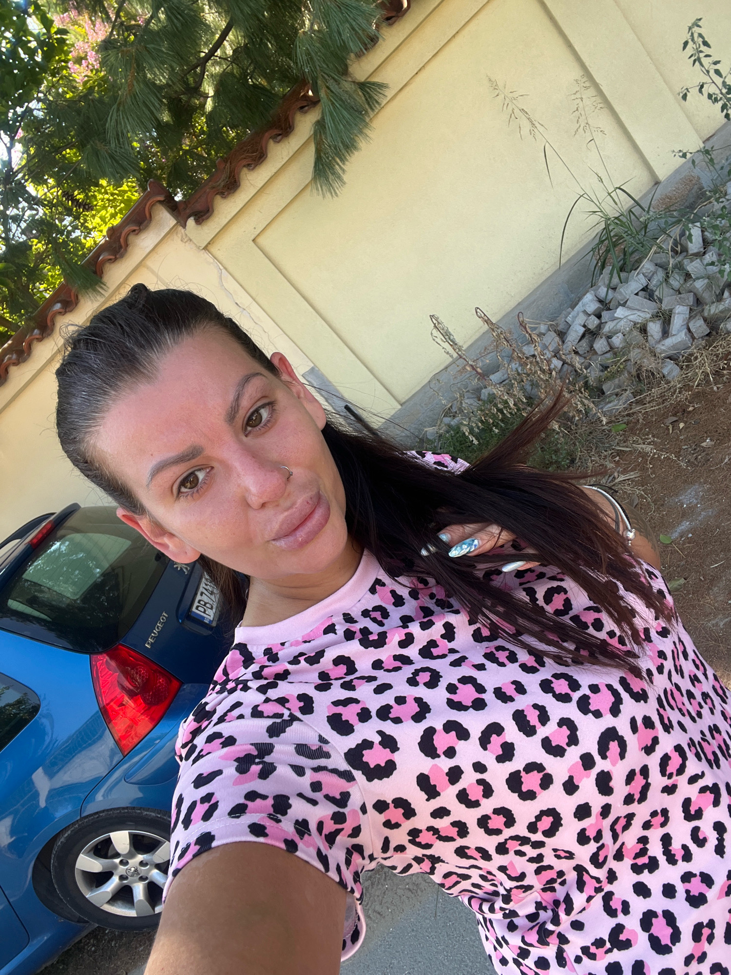 The image is a selfie taken outdoors, with a woman as the central subject. She has dark brown hair pulled back and is wearing a pink t-shirt with a black and pink leopard print. She has a nose ring and full lips. Her nails are long and decorated with a blue and silver glitter design. Behind her, a blue Peugeot car is visible, with its rear taillight and license plate showing. The environment appears to be a street or yard, with a pale yellow wall in the background, a tiled roof on a building, some dry vegetation, and a pile of grey bricks. The lighting suggests it is daytime. The license plate of the car is partially visible and appears to be Bulgarian, indicating the photo was likely taken in Bulgaria. Although the specific location within Plovdiv is not identifiable from the visual cues, the context points to an outdoor setting in the city.