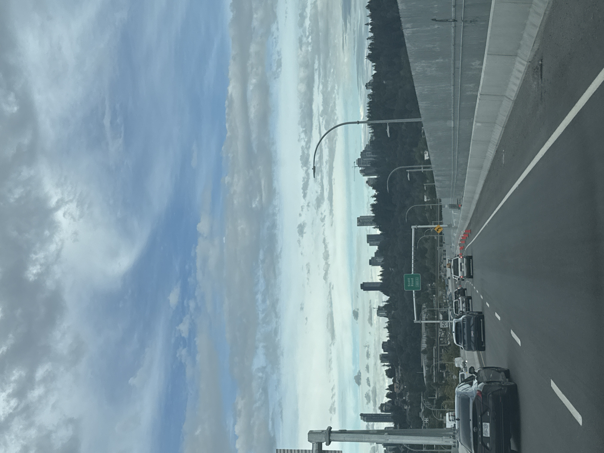The image captures a view from a vehicle traveling on a highway, likely in or near Surrey, Canada. The foreground shows a multi-lane highway with several cars driving. In the middle ground, a city skyline with numerous tall buildings and dense greenery is visible on a hillside. A green highway sign indicating "Scott Road" with a distance of "400 m" is present. The sky is filled with a mix of clouds, with some patches of blue. The overall scene depicts an urban landscape viewed from a commuting route.
