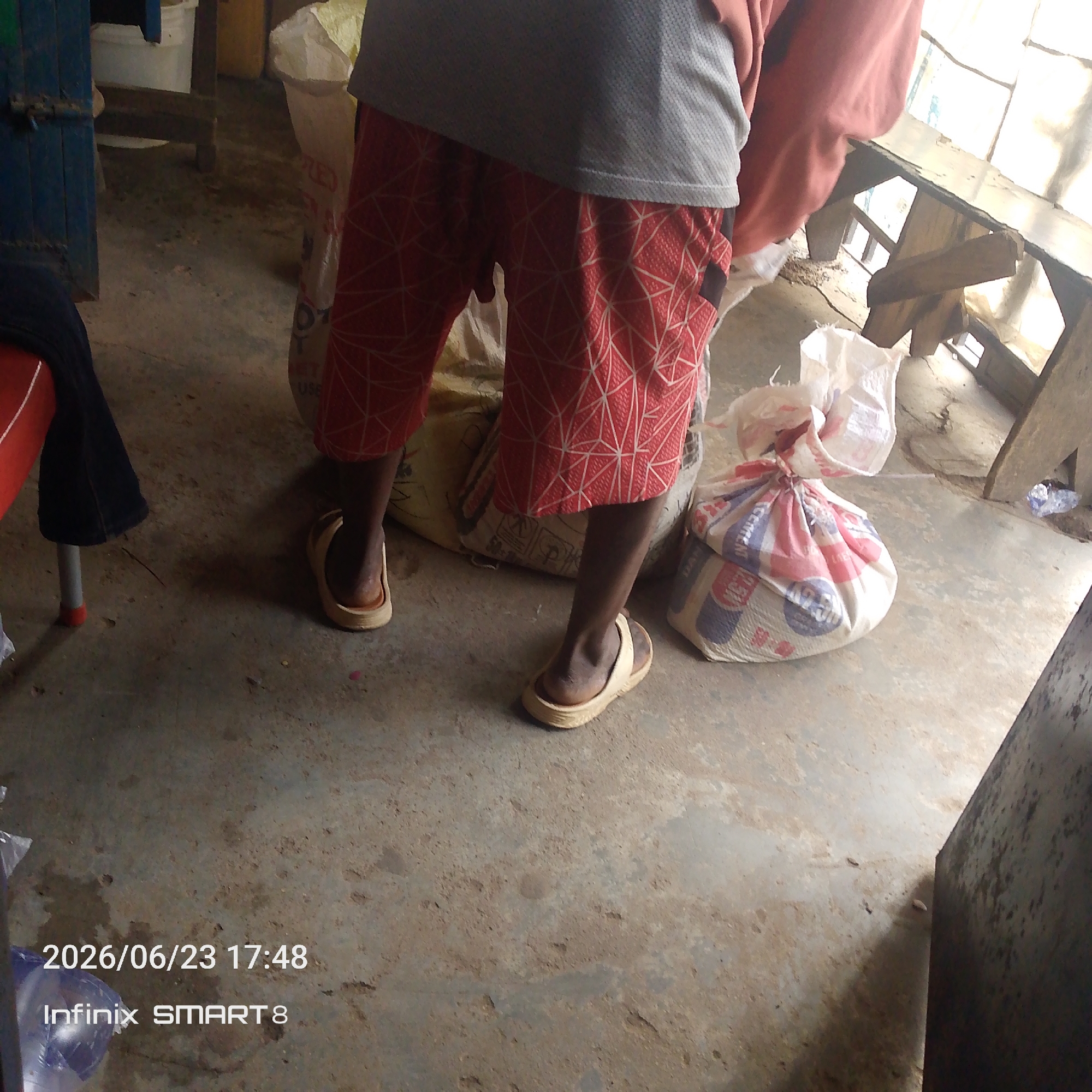 The image depicts a ground-level view of an indoor scene, likely a storage area or shop. A person, cropped from the waist down, is standing on a concrete floor. They are wearing red shorts with a white geometric pattern and light-colored sandals. The person is holding or standing near several large, tied sacks, one of which is white with red and blue printing. Another sack is visible behind the person, appearing to be a lighter color. To the left, a red chair with a dark blue cloth draped over it is partially visible. The background includes wooden structures, possibly shelving or a counter, and a white plastic bag or container. The overall environment appears somewhat cluttered and utilitarian. A date and time stamp, "2026/06/23 17:48", and a device name, "Infinix SMART 8", are overlaid on the bottom left of the image.
