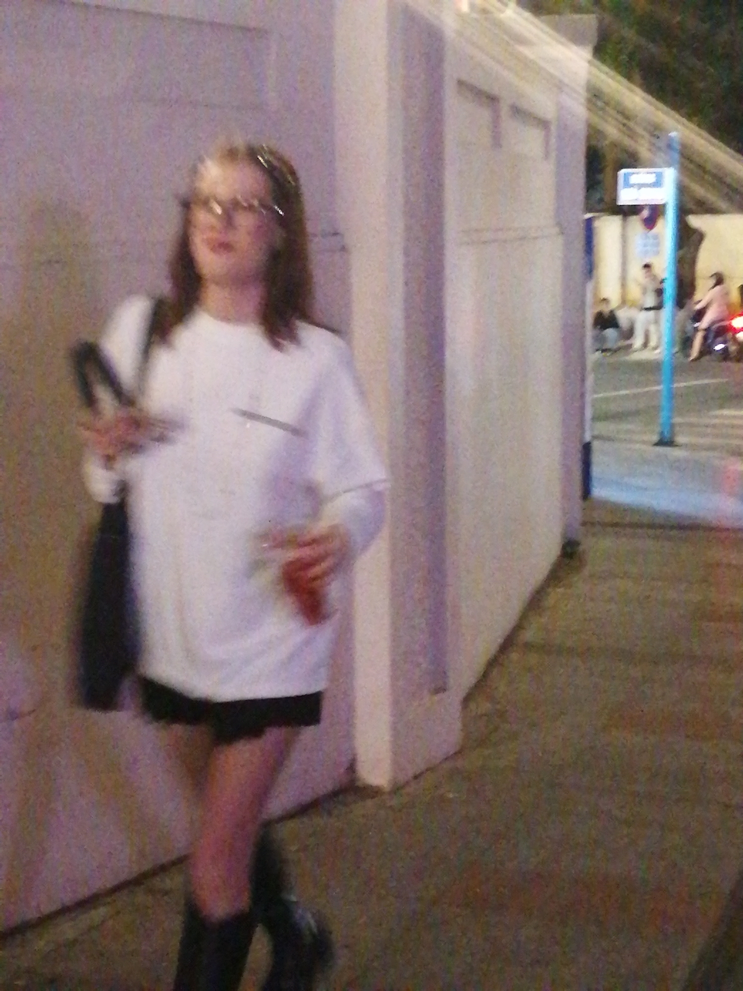 A person with long brown hair and glasses is walking, holding a dark bag and a drink. They are wearing a white long-sleeved shirt and black shorts, with dark boots. The person is in an urban setting, possibly a street or walkway, with a light-colored wall on the left and a sidewalk or road on the right. In the background, other people are visible, some sitting and others on a scooter. A blue pole with a white sign is also present, suggesting a public area. The lighting indicates it is likely evening or night. The overall scene appears to be a street in Da Lat, Vietnam.