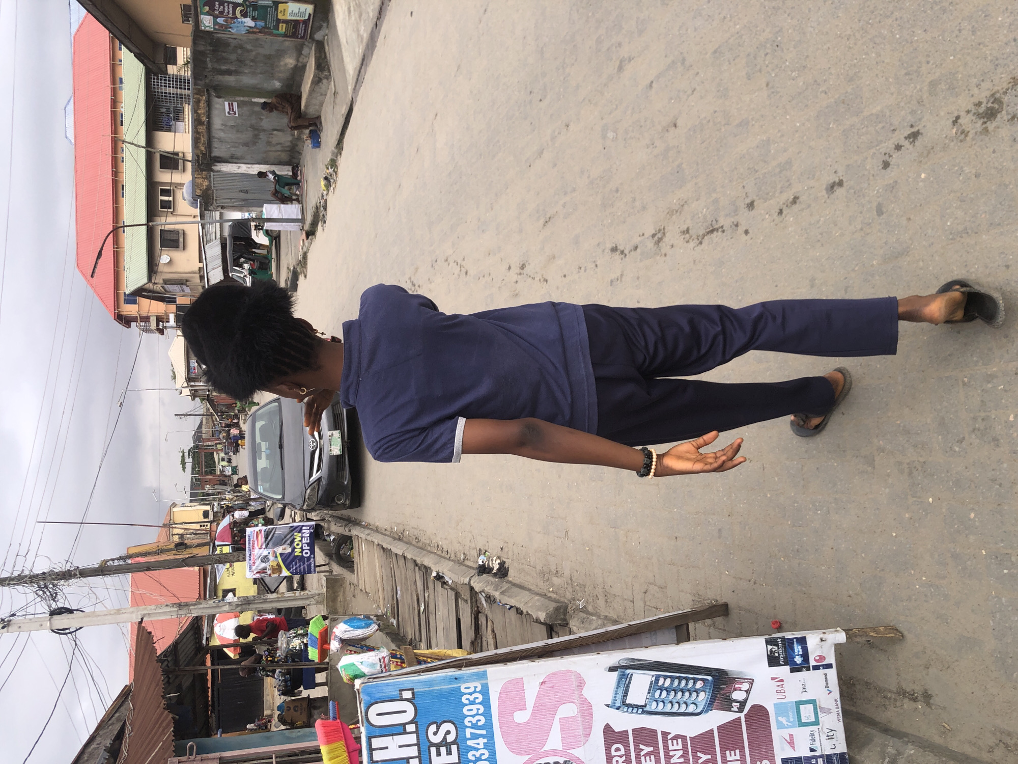 The image depicts a street scene in Ado, Nigeria. In the foreground, a young woman with braided hair is walking away from the camera. She is wearing a navy blue polo shirt and dark blue pants, with flip-flops on her feet. A beaded bracelet is visible on her left wrist. To the left of the woman, a white advertising board is propped up, displaying various logos of financial institutions and a phone number. Further down the street, a dark-colored car is parked. Several buildings line the street, some with corrugated iron roofs and others with more modern designs. A utility pole with numerous wires stands prominently on the left side of the road. The street itself appears to be paved with asphalt and shows signs of wear and tear, with debris scattered along the edges. The sky is overcast.