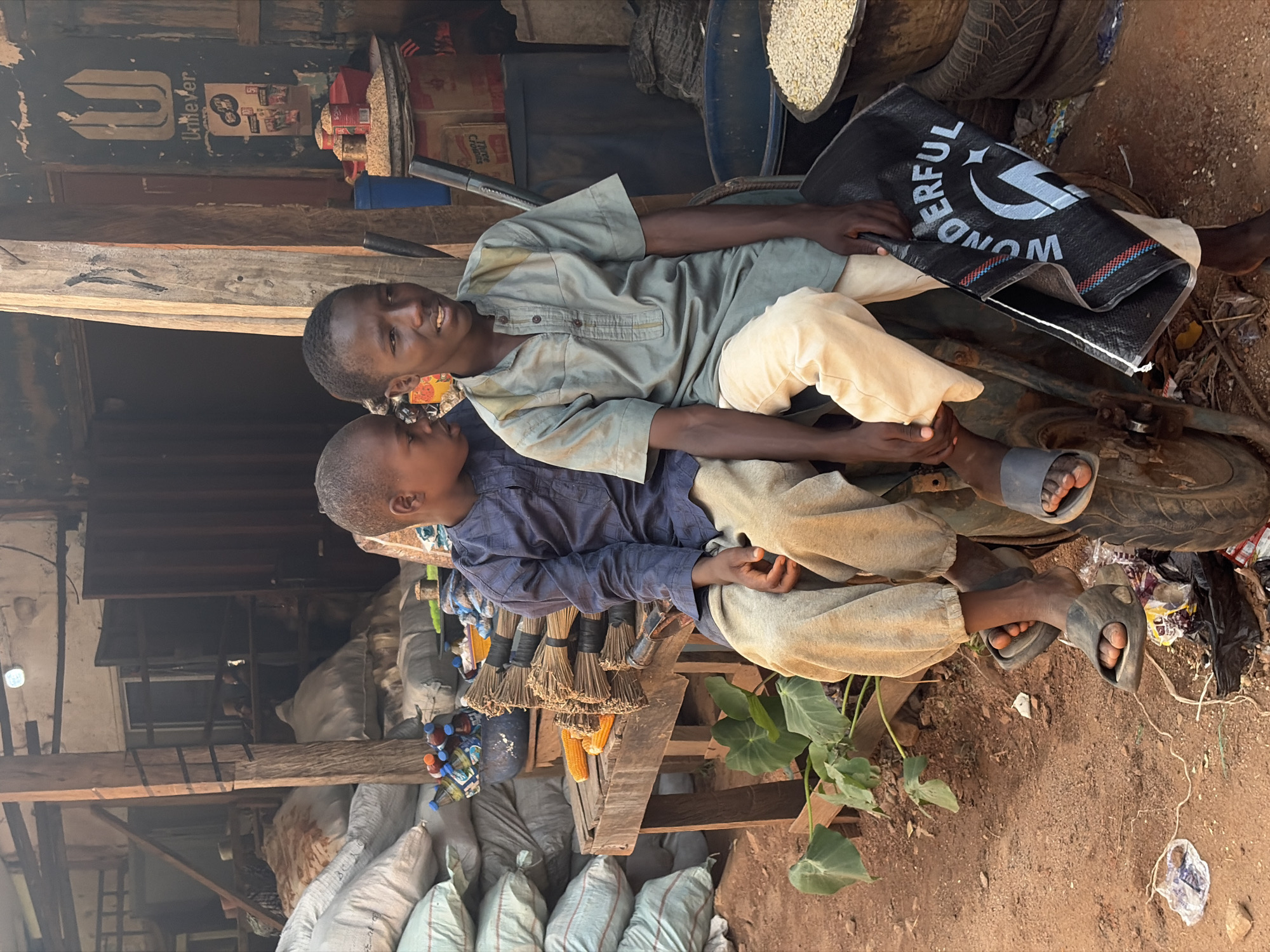 This media file features two young boys, likely teenagers, resting amidst an outdoor marketplace in Jos, Nigeria. The boy in the foreground, wearing a blue long-sleeved shirt and light-colored trousers, is looking towards the other boy. The second boy, wearing a pale green tunic and light-colored trousers, is looking towards the camera and appears to be smiling or talking. They are sitting on what looks like part of a vehicle, possibly a motorcycle or a cart, with a black woven bag labeled "WONDERFUL" with a stylized logo placed in front of them.
The setting is a bustling, somewhat rustic marketplace. Behind the boys, there are shelves stocked with various goods, including bags of grain or feed, cans, and other items, suggesting a place where produce and supplies are sold. Bundles of brooms and ears of corn are also visible, adding to the market atmosphere. In the background, there are stacked tires and more bags, indicating a storage or vendor area. The ground appears to be dirt, typical of many outdoor markets.
The lighting suggests it is daytime, with the sun casting shadows. The overall mood seems relaxed and informal, with the boys taking a break. There are no explicit indications of weather, but the scene appears to be dry. The presence of the "WONDERFUL" bag and the general market scene are key visual details. The specific location cues, like the types of goods and the building materials (wood and corrugated metal), are consistent with an environment found in Jos, Nigeria.