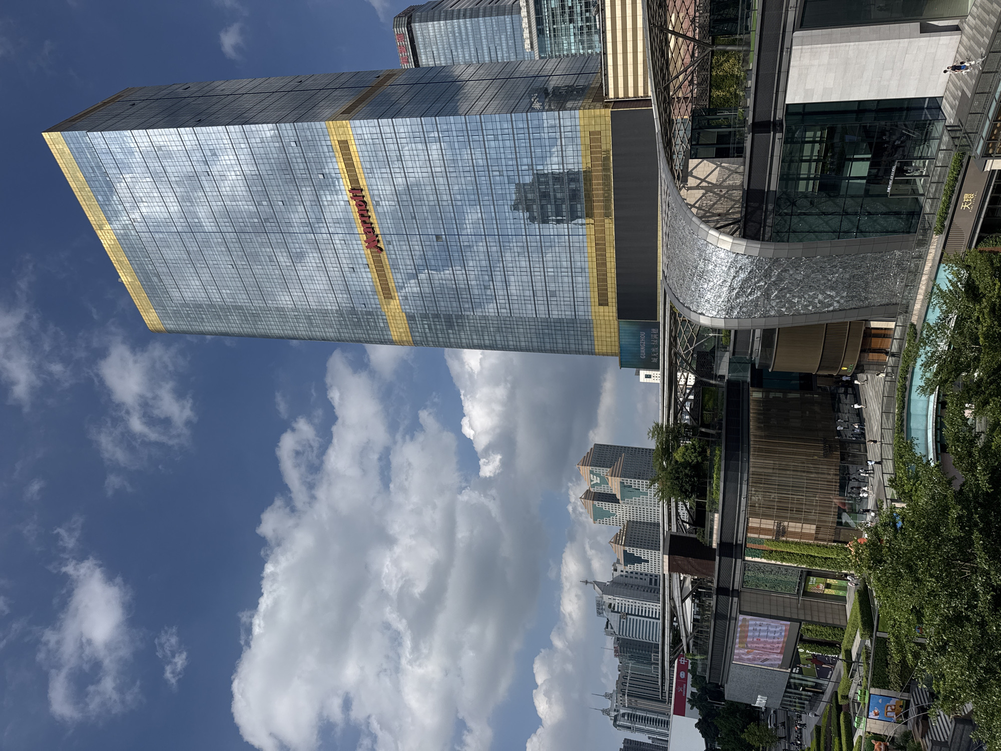 The image features a contemporary urban scene in the Tianhe District, China. A towering skyscraper with a reflective glass facade and prominent "Marriott" signage dominates the left side, reflecting the sky with scattered clouds. Gold-colored accents adorn the upper and lower sections of the building. Adjacent to the skyscraper is a modern architectural structure with a curved, textured surface, possibly part of a commercial complex. In the background, a cluster of high-rise buildings suggests a densely populated urban environment. At street level, greenery, trees, and pedestrian areas are visible. The image is taken at an angle, providing a view of the sky, the top of the buildings, and the streetscape below.