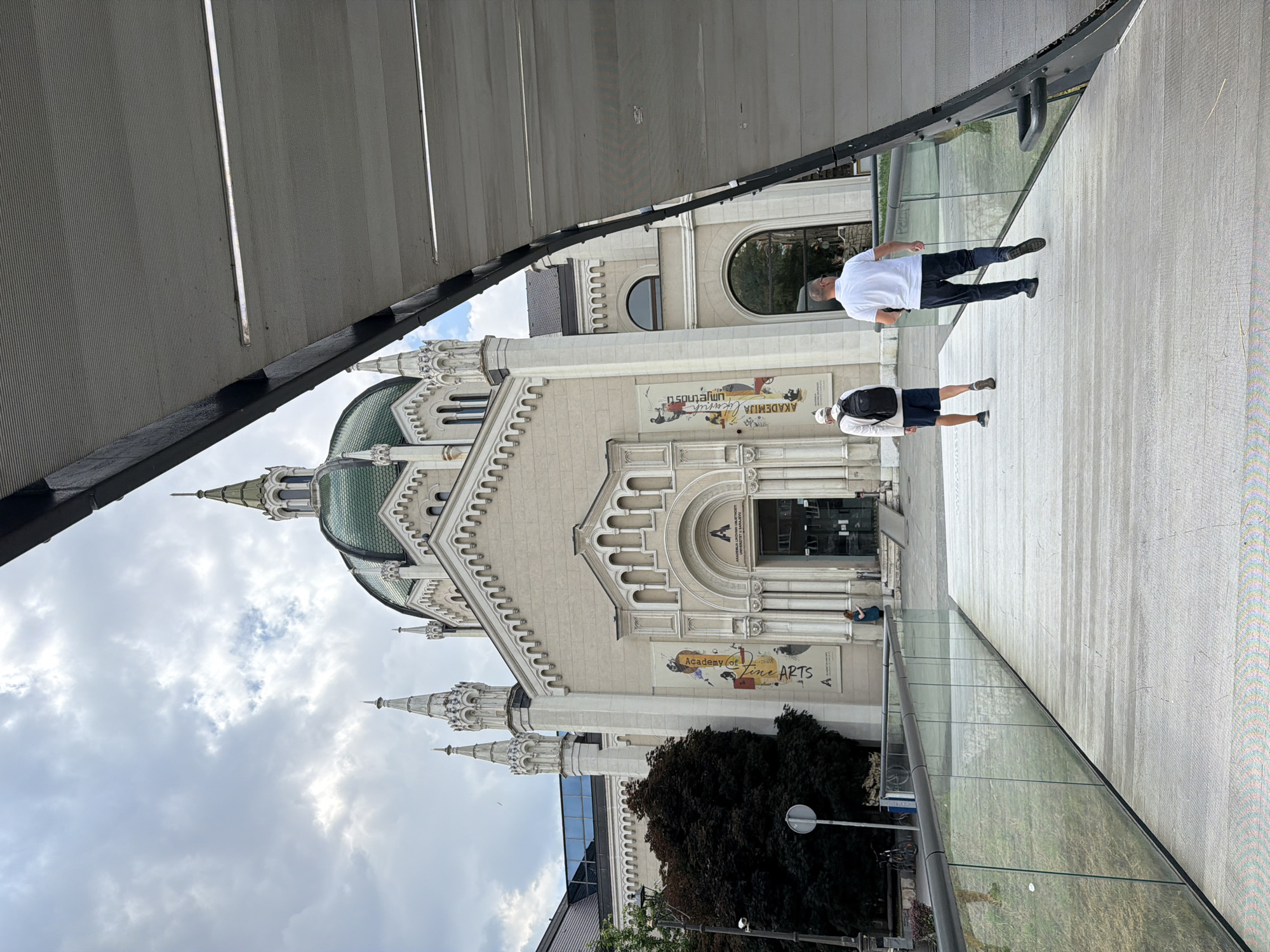 The image shows the Academy of Fine Arts building in Sarajevo, Bosnia and Herzegovina. Two men are walking on a modern elevated walkway towards the building. The building has a prominent green dome, white facade, and decorative spires. Banners with artwork and text are displayed on the building's exterior. The walkway is made of light-colored wood and has glass railings. The sky is overcast.