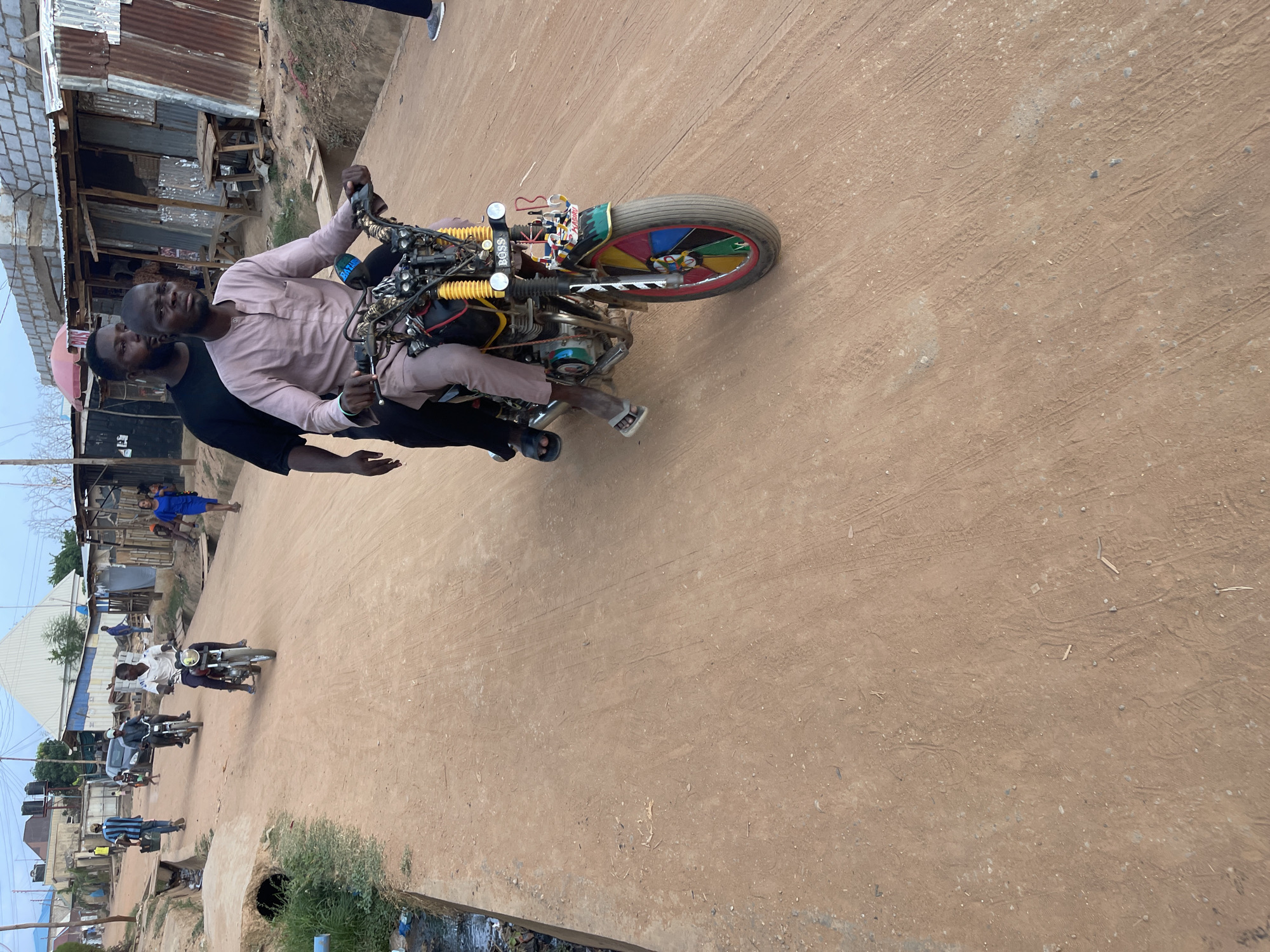 The image depicts two men riding a motorcycle on a dirt road in Karmajiji, Nigeria. The rider, wearing a light pink shirt and pants, is holding the handlebars. The passenger, wearing a black shirt, is behind him. The motorcycle has a decorated front wheel with multiple colors. The road is dirt and there are some buildings, people and motorcycles on the background. There is also a gutter with trash in the lower left part of the image.