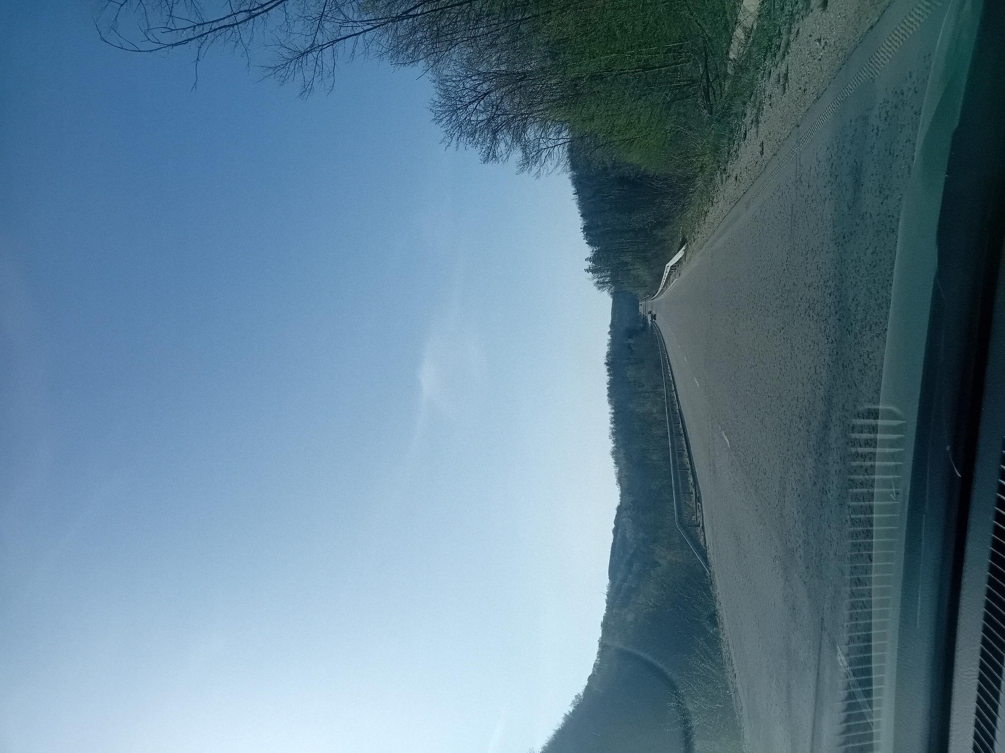 The image depicts a view from inside a vehicle, looking out onto a road. The road stretches into the distance, flanked by trees and vegetation on one side and a slope on the other. The sky above is clear and blue. The location is unknown.