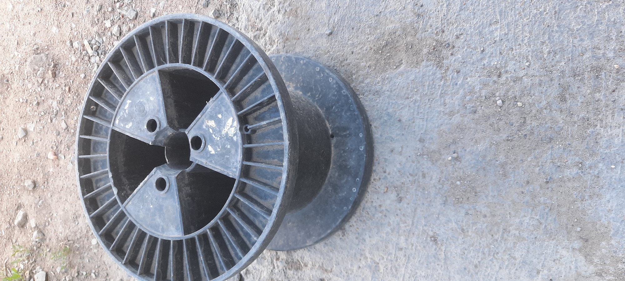 A black plastic cable spool sits on a concrete surface in an outdoor environment. The spool has a large central opening and spokes radiating outwards on its upper surface, with a wider base. The ground around the spool is a mixture of concrete and dirt with small stones. There is no visible damage or wear on the spool, except for some white scuff marks on the inner surfaces of the spokes. The background shows more dirt and stones. The image appears to be taken in Zhasaqan, Kazakhstan.