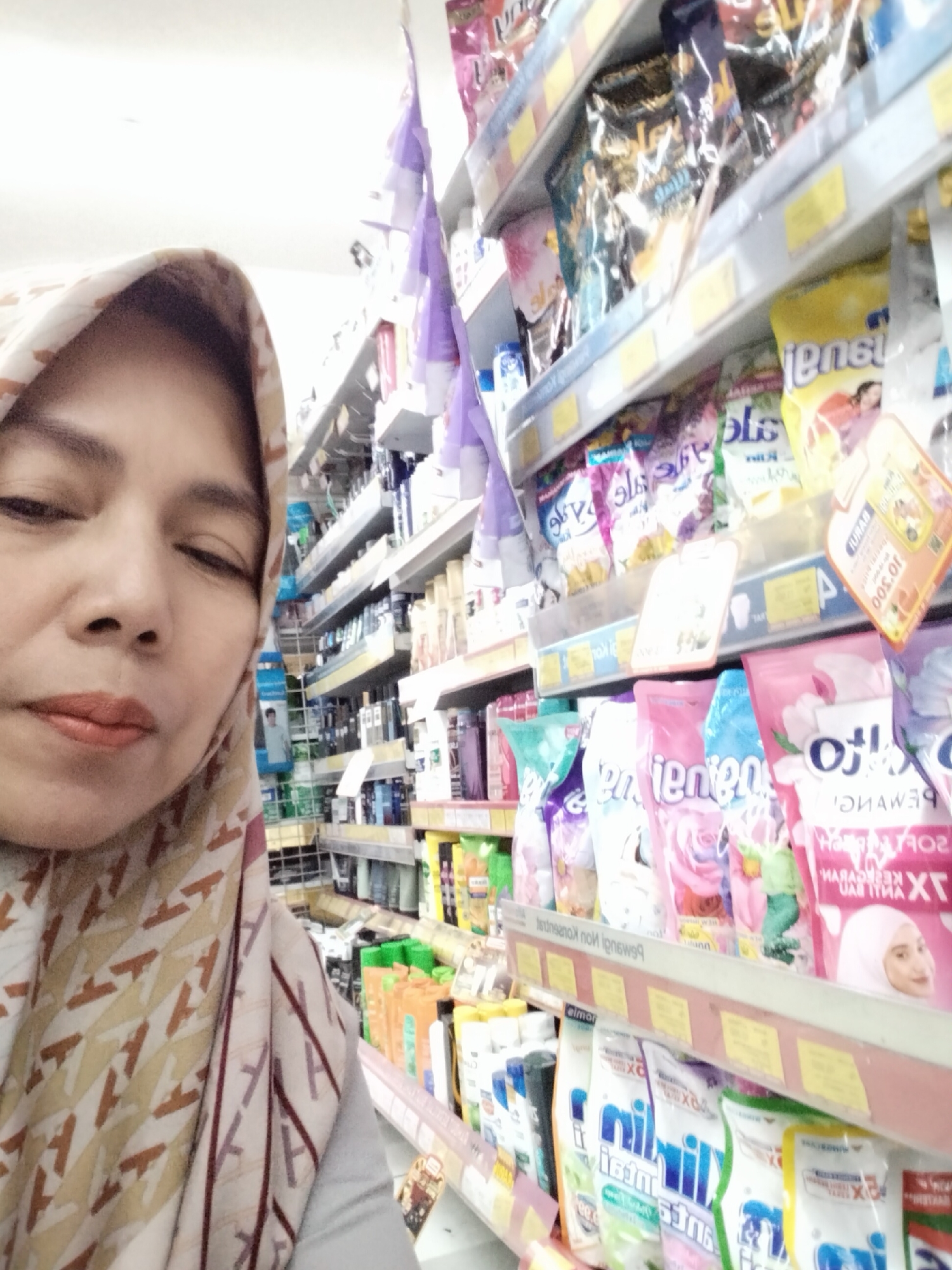A woman wearing a patterned hijab is taking a selfie in a brightly lit aisle of a store. She is positioned in the foreground, partially obscured by the fabric of her hijab. The background reveals shelves stocked with various products, including detergent packets and bottles. The lighting is overhead and appears to be fluorescent, casting a cool tone on the scene. The focus is primarily on the woman's face and the immediate products on the shelves.