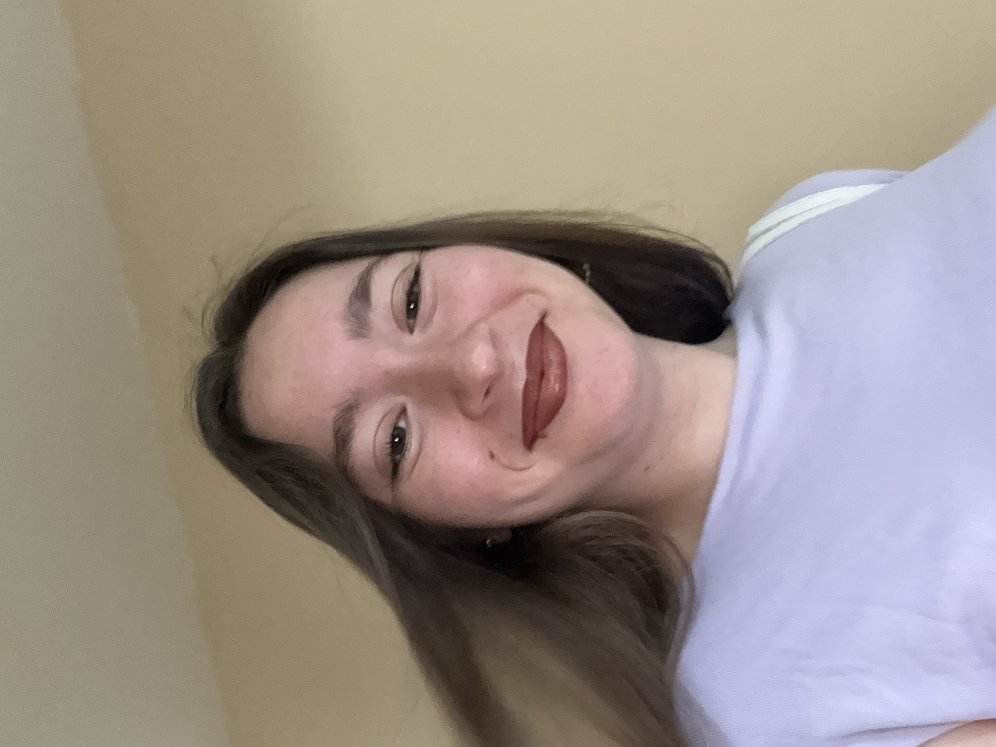 A young woman with long, dark brown hair is looking at the camera and smiling. She is wearing a light purple t-shirt and has on dark brown lipstick. Her hair is partially obscuring her face on the left side. The background is a plain, light brown wall. The lighting suggests an indoor environment. There is no identifiable information about the location in Sofia, Bulgaria.