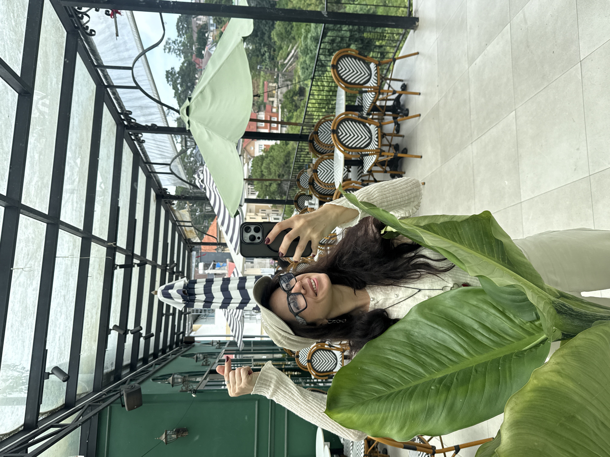 A woman is taking a selfie on an outdoor patio in Da Lat, Vietnam. She is wearing a beige cap, glasses, and a cream-colored sweater. She is holding a large green leaf in front of her and a smartphone in her right hand. The patio has tables and chairs with black and white striped cushions, and umbrellas. In the background, there are buildings and lush green hills, characteristic of Da Lat's landscape. The woman is smiling and giving a thumbs up. The setting appears to be a cafe or restaurant with a scenic view.