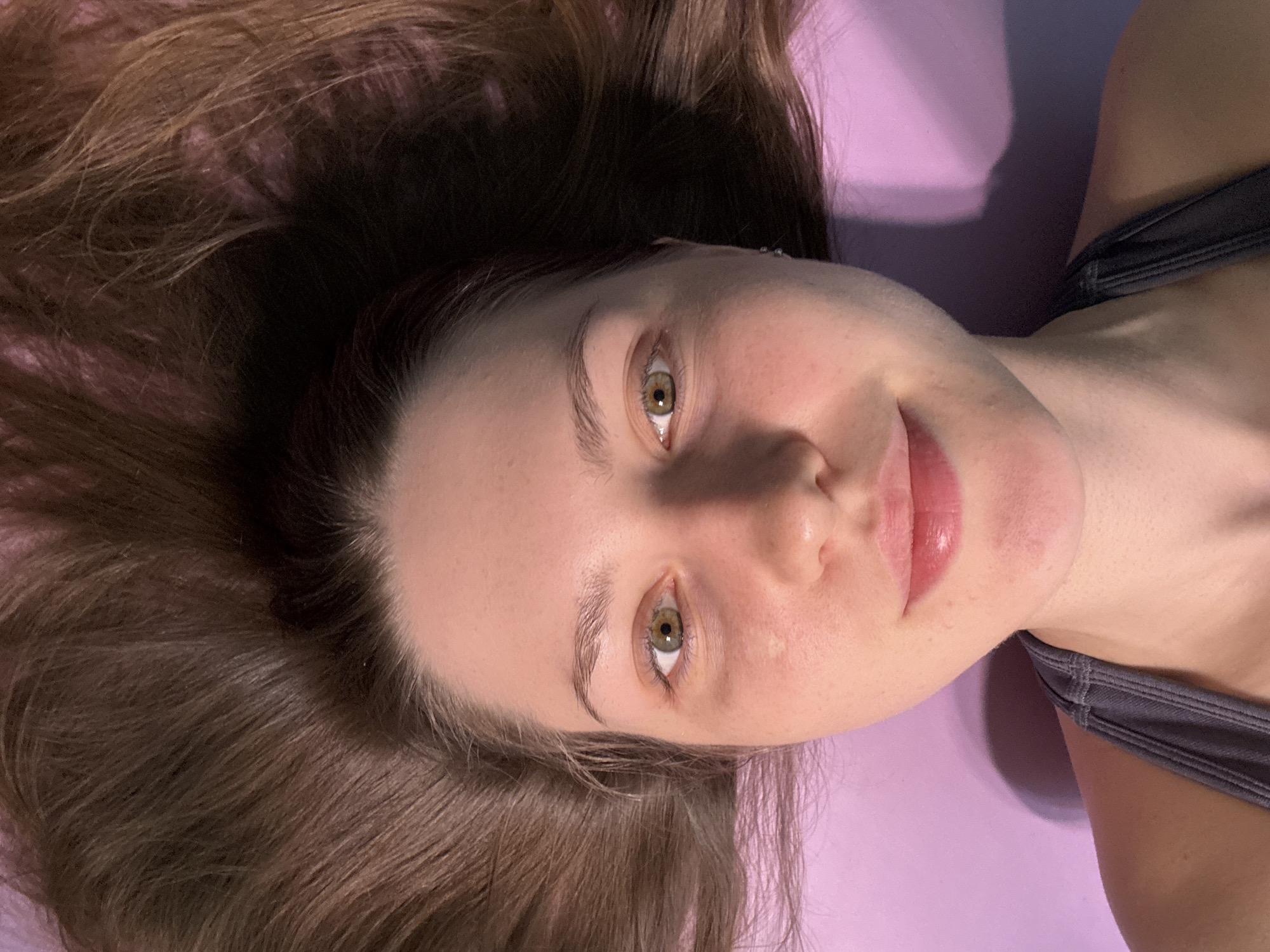 A close-up portrait shows a young woman with fair skin, brown hair, and striking hazel or green eyes, lying down and looking directly at the camera. Her long, brown hair is spread out around her head. She wears a dark grey top, possibly a sports bra or athletic wear. The woman displays a gentle, soft smile and a serene expression, with her gaze fixed on the viewer. The background is a uniform light purple or lavender surface, indicating an indoor setting, perhaps a mat or bed. The lighting appears to be artificial, creating soft shadows on the purple surface, particularly behind her head. There are no visible location cues within the image itself, though the context provided states the location is Saint Petersburg, Russia. No text is visible in the media file. The overall impression is one of calm and natural composure.