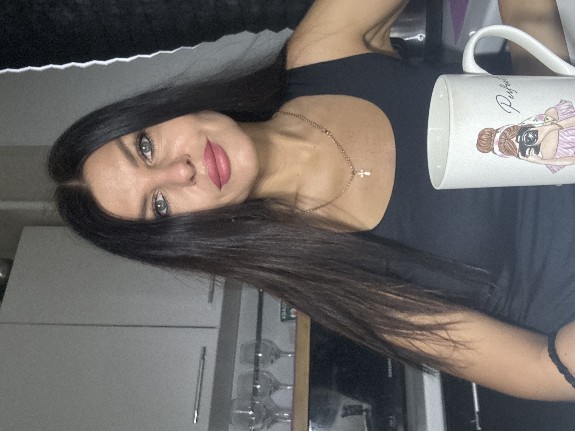 A portrait-oriented image, rotated 90 degrees counter-clockwise, features a woman looking directly at the camera while holding a mug in an indoor setting. The woman has long, dark hair, parted down the middle, and striking light-colored eyes, possibly blue or green. She wears pink lipstick and is dressed in a black sleeveless top. A delicate gold-colored chain with a small cross pendant rests on her collarbone. Her left arm is visible, adorned with a black band or hair tie on her wrist.
She holds a white ceramic mug, which displays an illustration of a woman with her hair in a bun, wearing a pink outfit, and holding a camera. The word "Perfect" is written in a decorative script above the illustration.
The background suggests a domestic environment, likely a kitchen or a living space with kitchen features. Light-colored (white or light gray) cabinets are visible, and below them, several wine glasses are hanging upside down from a rack. A dark appliance, possibly a microwave or oven, can also be seen. A blurred black textured surface occupies the top portion of the frame, possibly part of a piece of furniture. The lighting is soft and consistent with indoor artificial light. The woman maintains a calm or slightly smiling expression. The location context for this image is Chaikovsky, Russia.