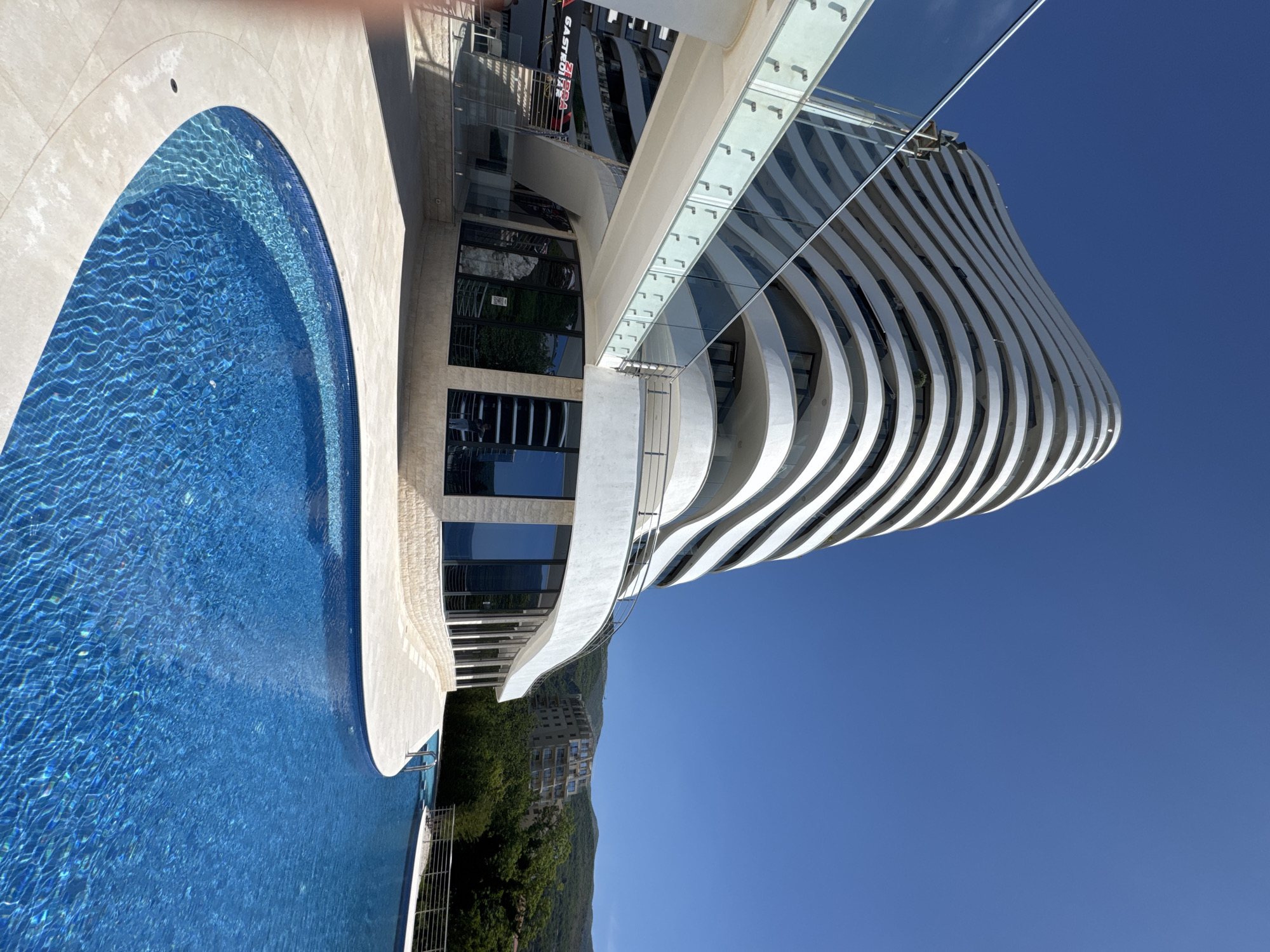 The image depicts a modern architectural building in Boreti, Montenegro, adjacent to a blue-tiled swimming pool. The building's design is characterized by curved, layered white balconies, giving it a distinctive, undulating facade. A glass-enclosed walkway connects different sections of the building. The lower part of the building features vertical windows and stone-like textures. In the background, a green hillside dotted with structures can be seen under a clear, bright blue sky.