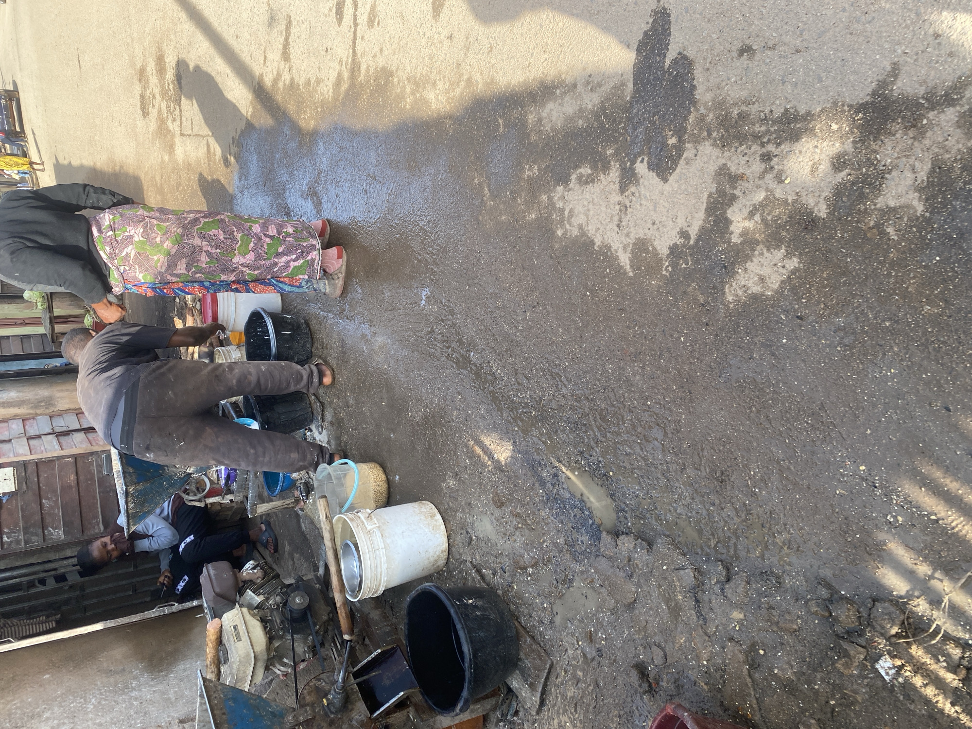 In a street scene in Jos, Nigeria, a young woman sits in the shade of a building, looking at her phone. She is dressed in casual wear, including dark pants and a grey hoodie. Beside her, a man in dark pants and a long-sleeved shirt is engaged in some activity involving a water bucket and a food processor. He is wearing a colorful skirt-like garment over his pants. In the foreground, several buckets are visible, some containing water and other materials. The ground is wet and uneven, suggesting recent water activity. The background shows more buildings and a glimpse of traffic on the street. The overall impression is one of everyday life and work in a bustling urban environment.